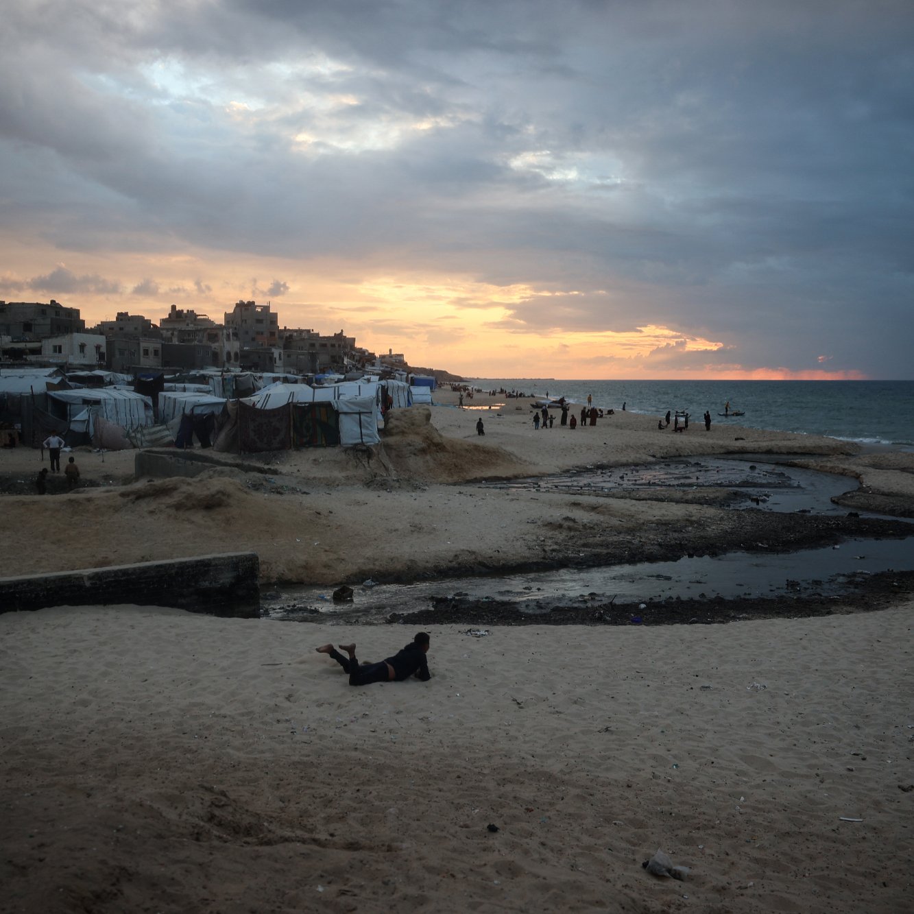 L'image montre une plage à l'heure du coucher de soleil, avec des nuages sombres dans le ciel. Au premier plan, on aperçoit du sable et une petite étendue d'eau stagnante. On peut également voir des tentes ou des abris, probablement des habitations temporaires, ainsi que quelques personnes se déplaçant sur la plage. L'atmosphère est à la fois paisible et mélancolique, avec des teintes orange et grises dans le ciel.
