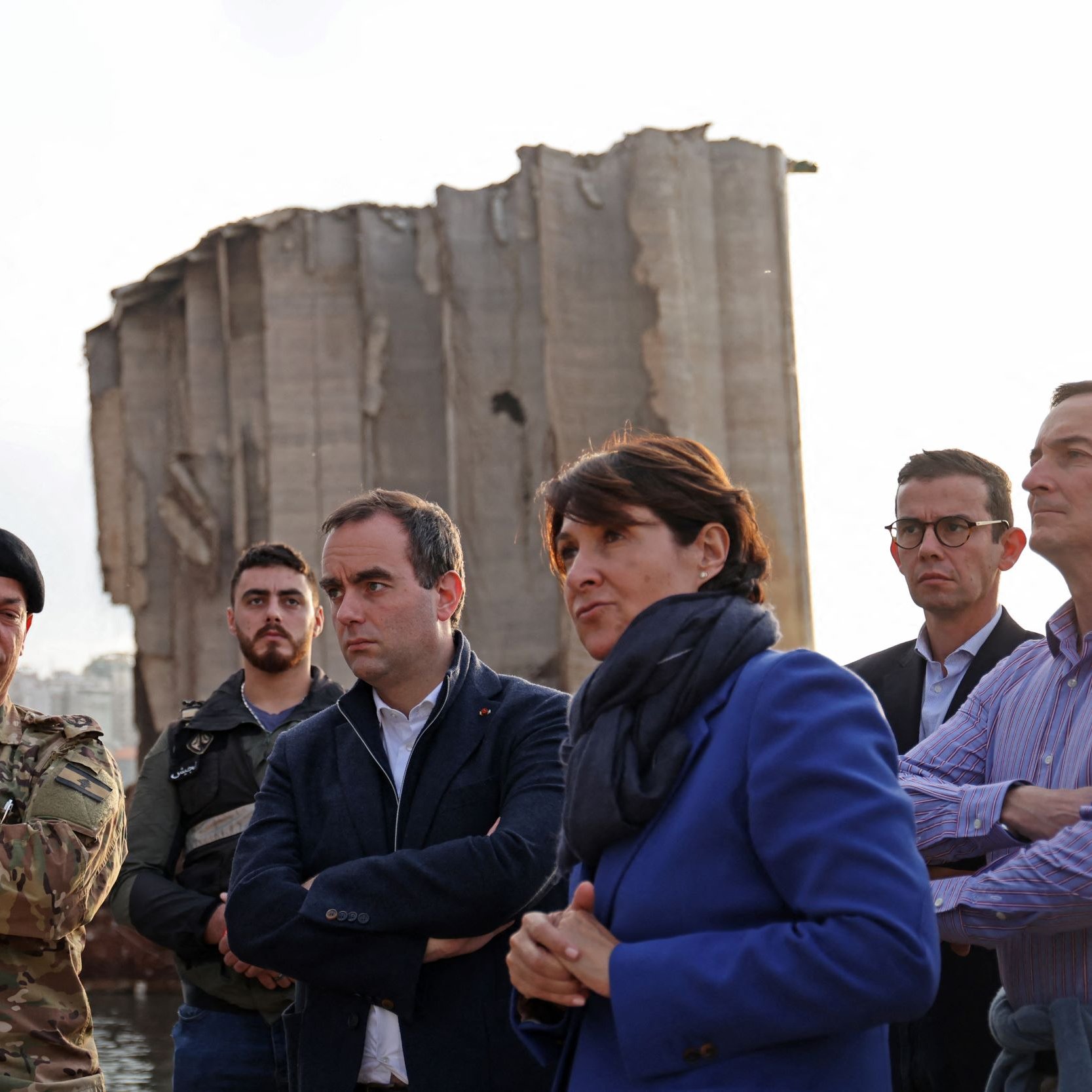 در این تصویر، گروهی از افراد در کنار یک ساختمان ویران، که ظاهراً بخشی از آن تخریب شده، ایستاده‌اند. یک نظامی با لباس نظامی و کلاه فرماندهی در سمت چپ قرار دارد. کنار او مردان و زنانی با لباس‌های رسمی و غیررسمی ایستاده‌اند و به نظر می‌رسد که در حال صحبت و تبادل نظر هستند. پس‌زمینه تصویر نشان‌دهنده شهری با ساختمان‌های بلند است. نور در تصویر کم است و فضای کلی حالت جدی و تاملی دارد.