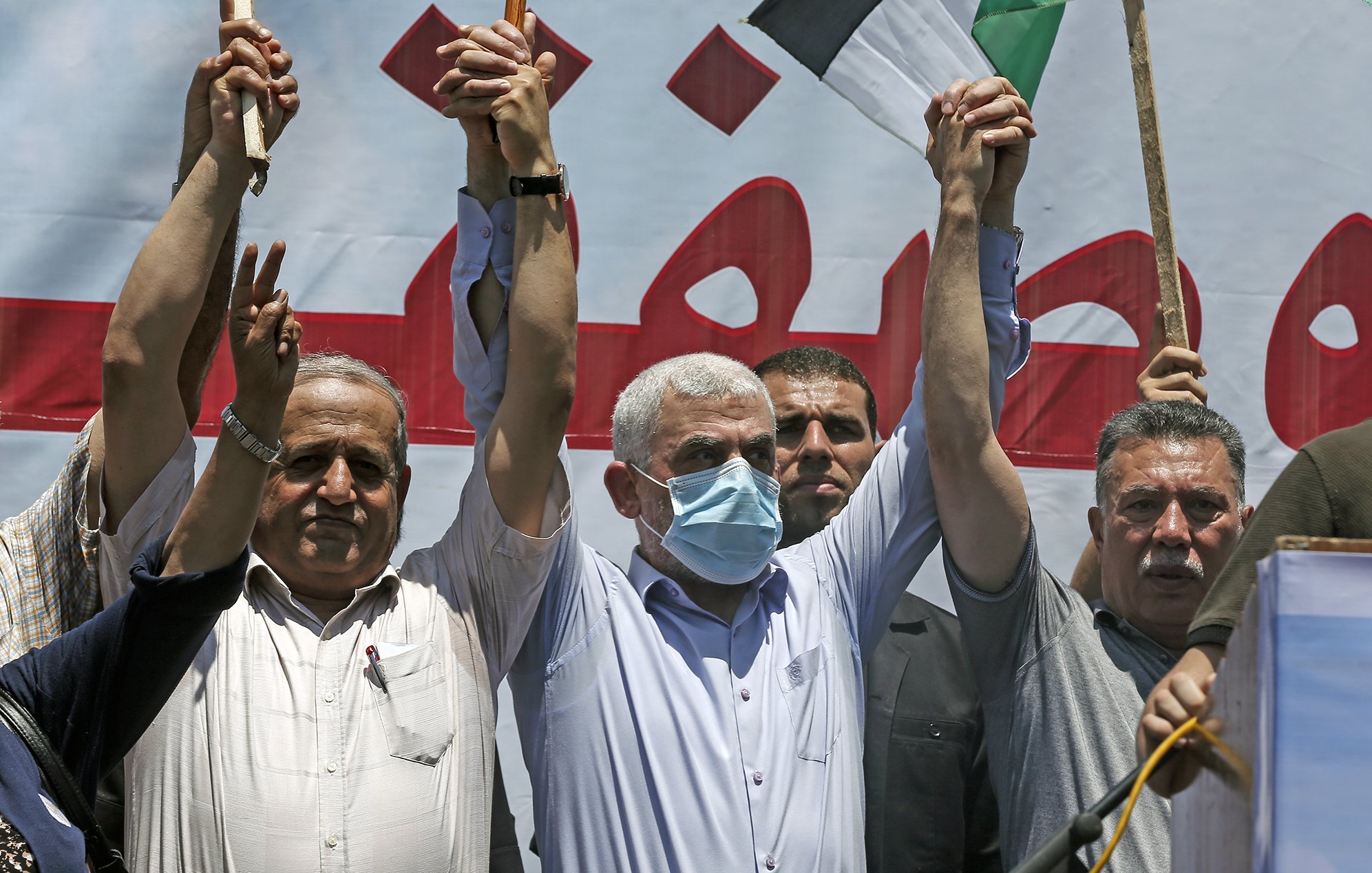 The image depicts a group of men at a public gathering or protest. They are raising their hands in the air, with some holding sticks or flags. The man at the center is wearing a mask and appears to be leading or participating actively in the event. In the background, there is a large banner in Arabic, suggesting a political or social message. The mood of the event seems to be one of solidarity or protest, with participants visibly engaged.