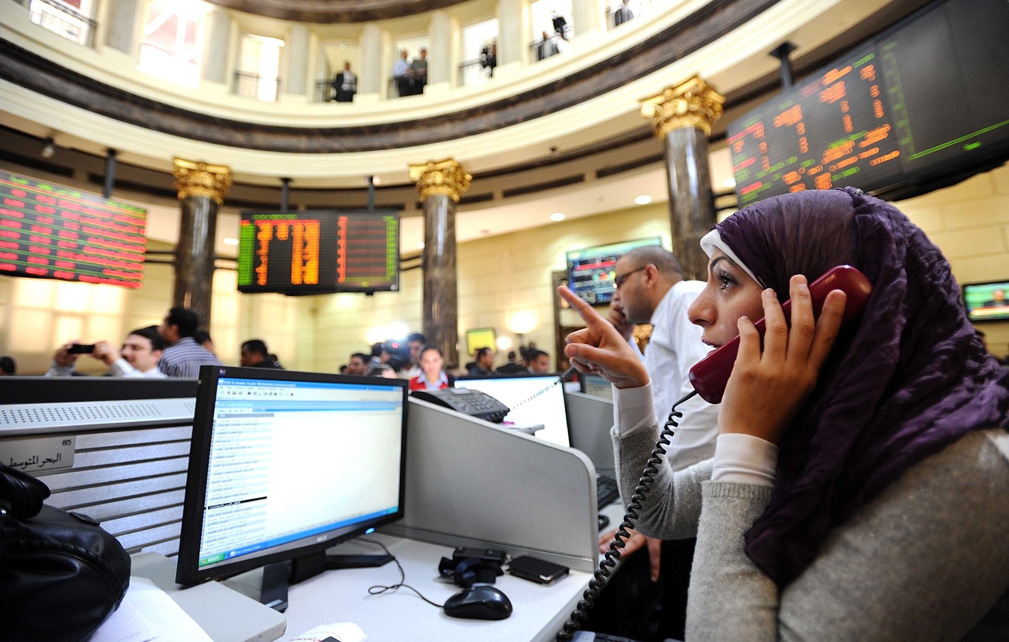 L'image montre une bourse avec plusieurs personnes en train de communiquer, probablement en train de négocier des actions. Au premier plan, une femme portant un hijab est concentrée sur un téléphone, avec un ordinateur devant elle affichant des données financières. En arrière-plan, on peut voir des écrans affichant des chiffres et des informations boursières, ainsi qu'un groupe de personnes observant attentivement la situation. L'ambiance semble dynamique et chargée d'activité.