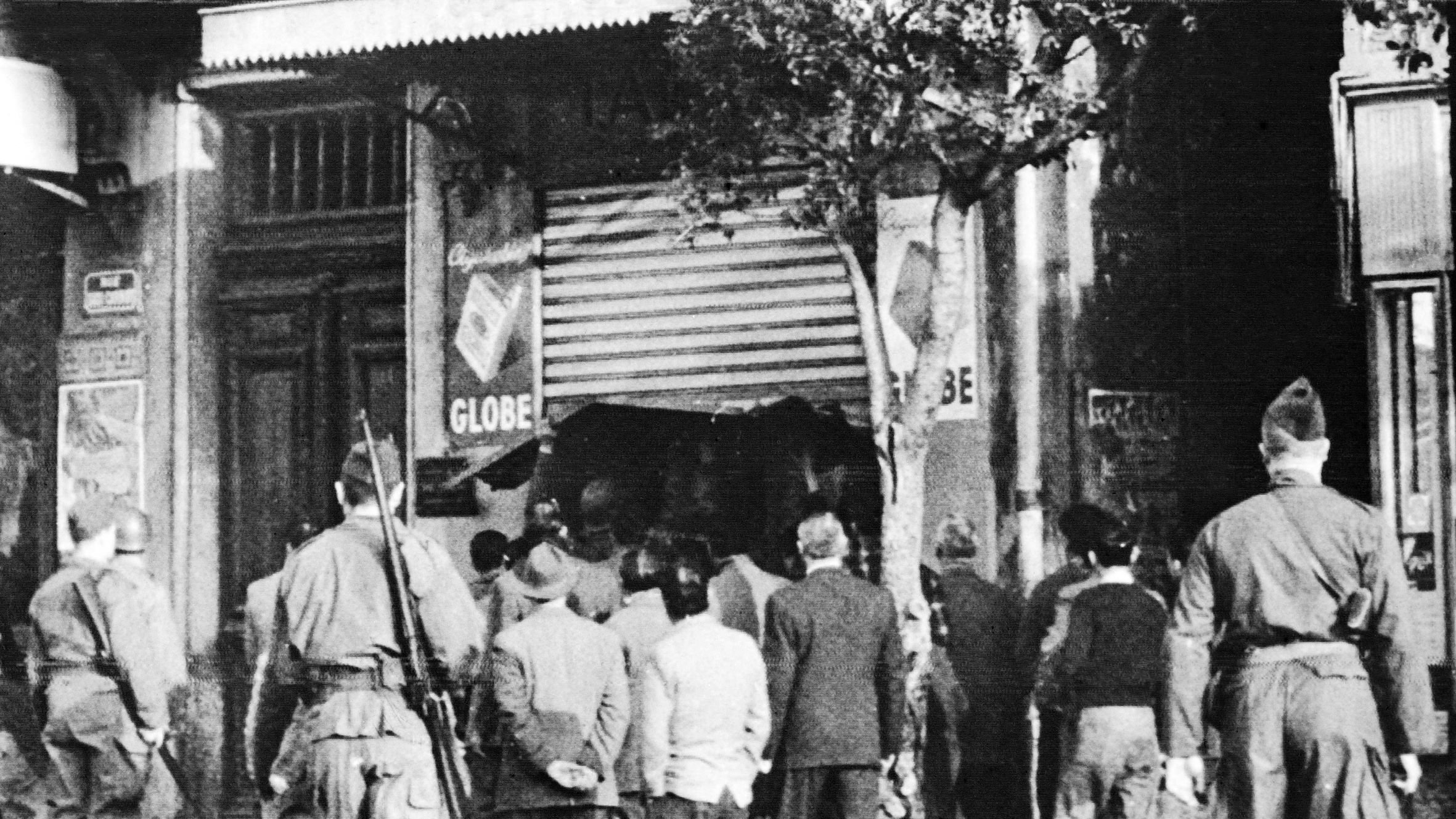 در این تصویر، گروهی از مردم در خیابان در حال نظارت به یک مکان هستند. تعدادی سرباز در نزدیکی آن‌ها ایستاده‌اند که به نظر می‌رسد در حال کنترل اوضاع هستند. این تصویر احتمالاً از یک زمان تاریخی گرفته شده است و نشان‌دهنده تنش یا رویدادی خاص در آن زمان است. حالت افراد و لباس‌هایشان به این احساس دامن می‌زند. برخی از افراد به سمت مغازه‌ای در حال حرکت هستند، در حالی که دیگران از دور نظاره‌گر هستند.