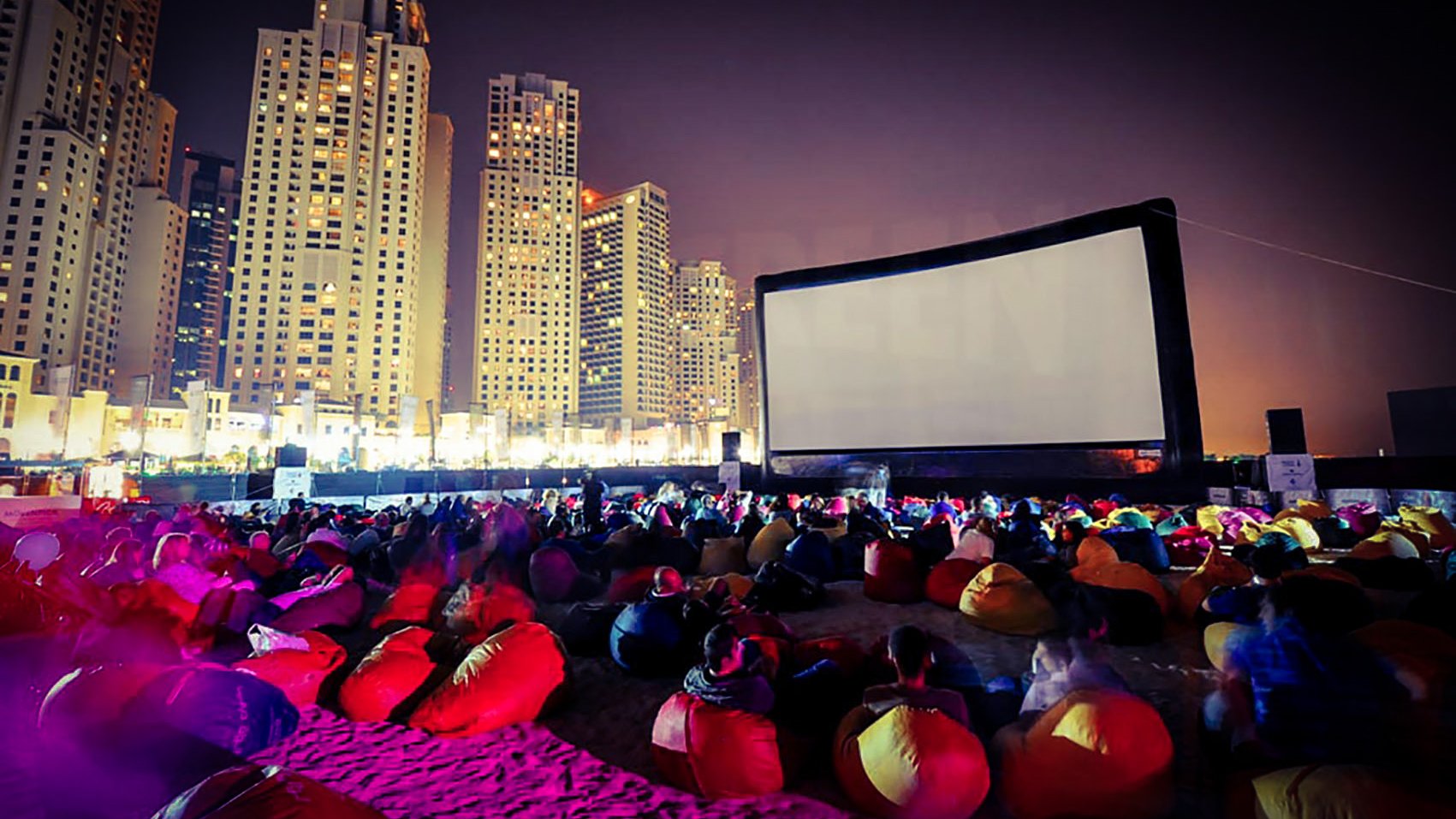 L'image montre une scène de cinéma en plein air sur une plage, animée pendant la nuit. Un grand écran est installé, et des groupes de personnes s'asseyent sur des poufs colorés, principalement de couleur rouge. En arrière-plan, on peut voir des gratte-ciels illuminés, créant une atmosphère vibrante et moderne. L'éclairage doux et les ombres ajoutent à l'ambiance festive du lieu.