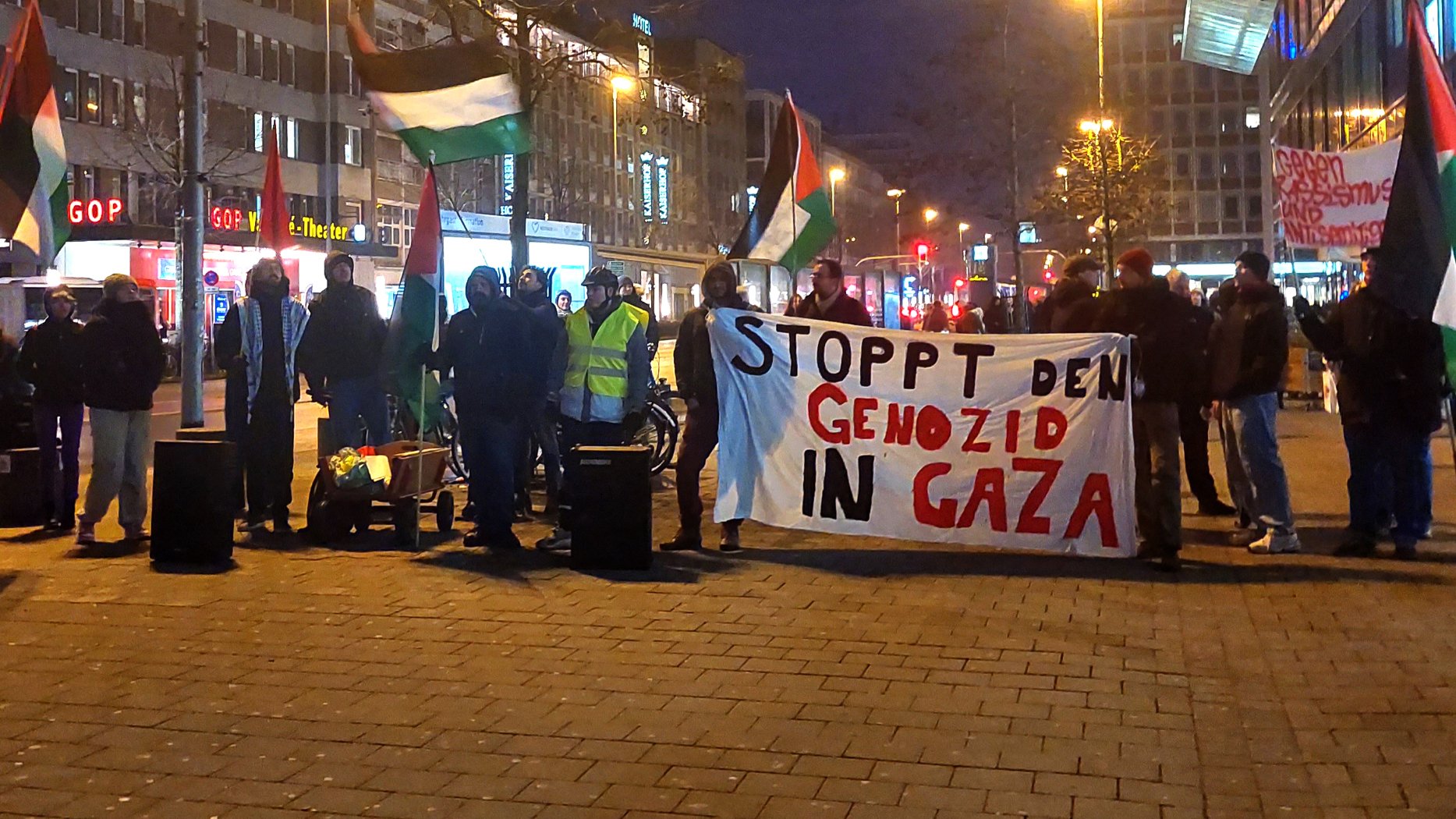 این تصویر مربوط به تجمعی است که در آن تعداد زیادی از افراد با پرچم‌های فلسطینی و یک banner بزرگ حضور دارند. روی بنر نوشته شده است: "STOPPT DEN GENOZID IN GAZA" که به معنای "نسل‌کشی در غزه را متوقف کنید" است. فضای تجمع شهری به نظر می‌رسد و شب است.