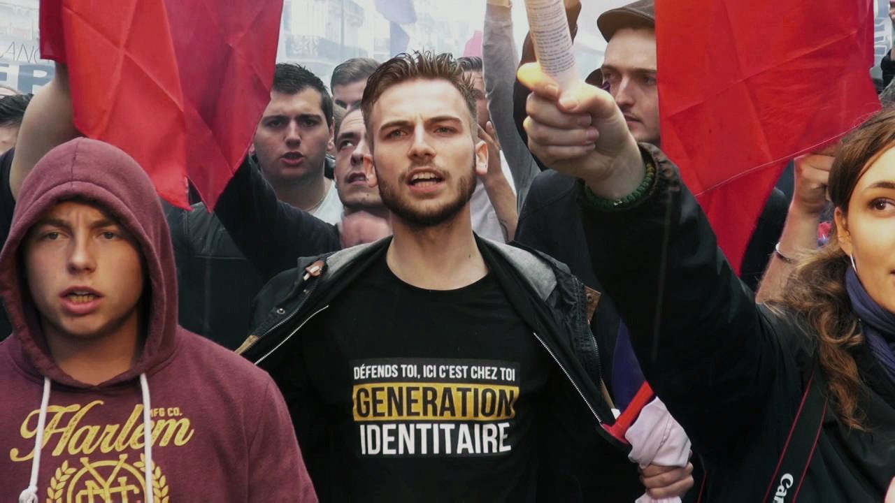 The image depicts a protest or demonstration scene. In the foreground, a young man stands prominently, wearing a black shirt with the text "GÉNÉRATION IDENTITAIRE" printed in yellow. He appears to be leading the group, holding a piece of paper in one hand and raising it in the air. Surrounding him are several other individuals, some of whom are holding red flags. The atmosphere seems charged and passionate, with many faces visible, indicative of a collective movement. The overall context suggests a rally focused on identity or nationalist themes.