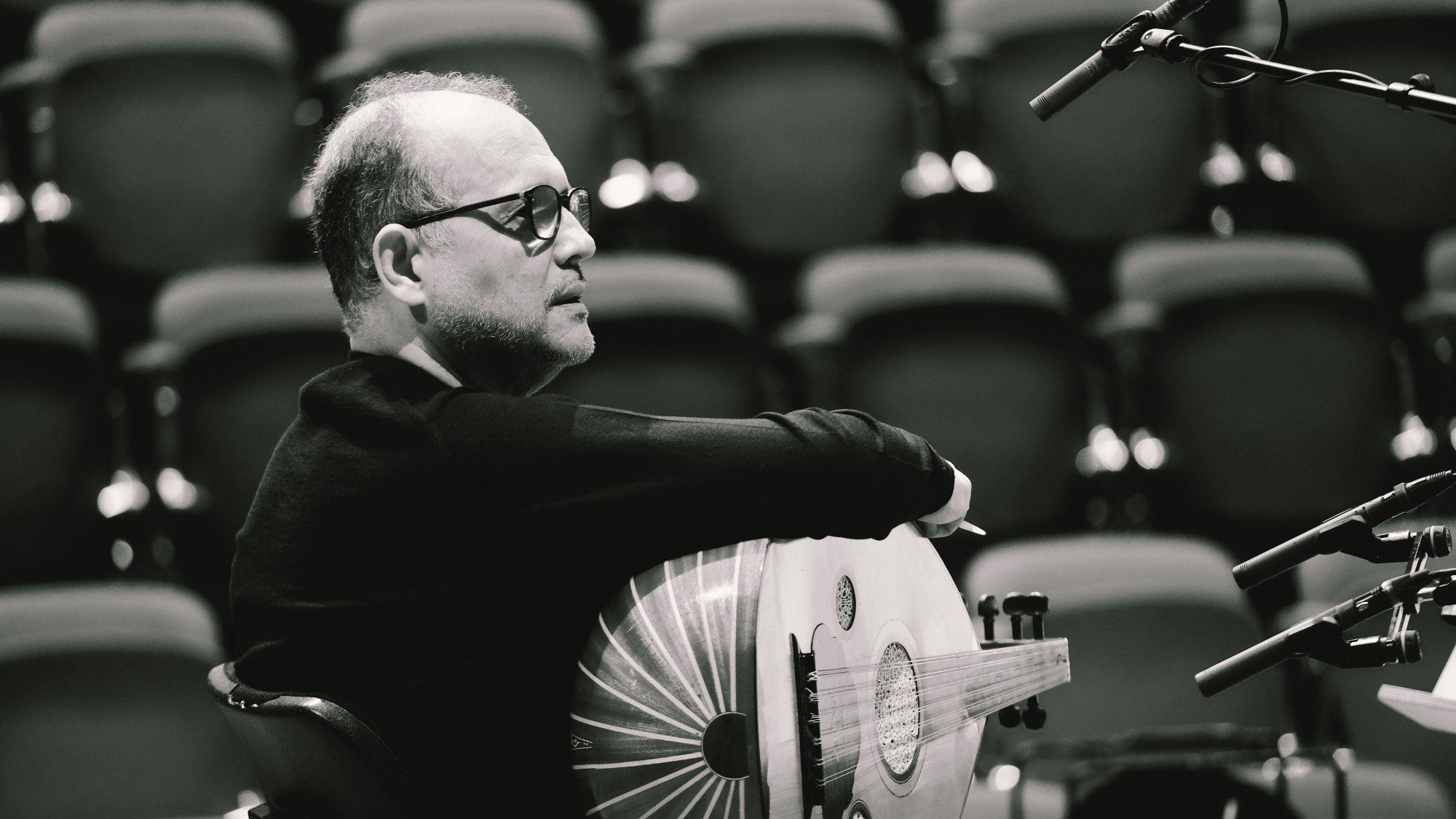 L'image montre un homme assis, tourné légèrement sur le côté. Il porte des lunettes et un pull noir. Dans ses bras, il tient un instrument de musique en forme de luth. L'arrière-plan est flou avec des rangées de sièges.