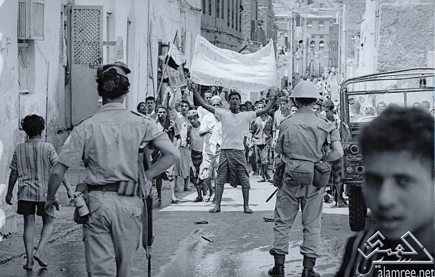 The image depicts a historical scene in a narrow street, likely during a protest or demonstration. In the foreground, a group of people is visible, with a man raising a banner or flag, indicating a spirited display. There are several individuals, including children, observing the scene. Two uniformed officers can be seen in the foreground, suggesting a presence of law enforcement. The atmosphere appears tense, with people engaged in an active moment, highlighting social or political unrest. The black-and-white format emphasizes the historical context of the image.