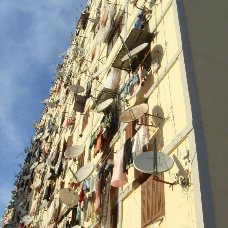 L'image montre un bâtiment d'habitation avec de nombreux balcons. Les murs de l'immeuble sont couverts de vêtements suspendus à sécher, créant un décor coloré et animé. De plus, plusieurs antennes parabolique sont fixées sur les façades, indiquant que les habitants ont accès à la télévision satellite. Le ciel est clair, ajoutant une lumière naturelle à la scène. L'ensemble donne une impression de vie communautaire et de simplicité dans un cadre urbain.