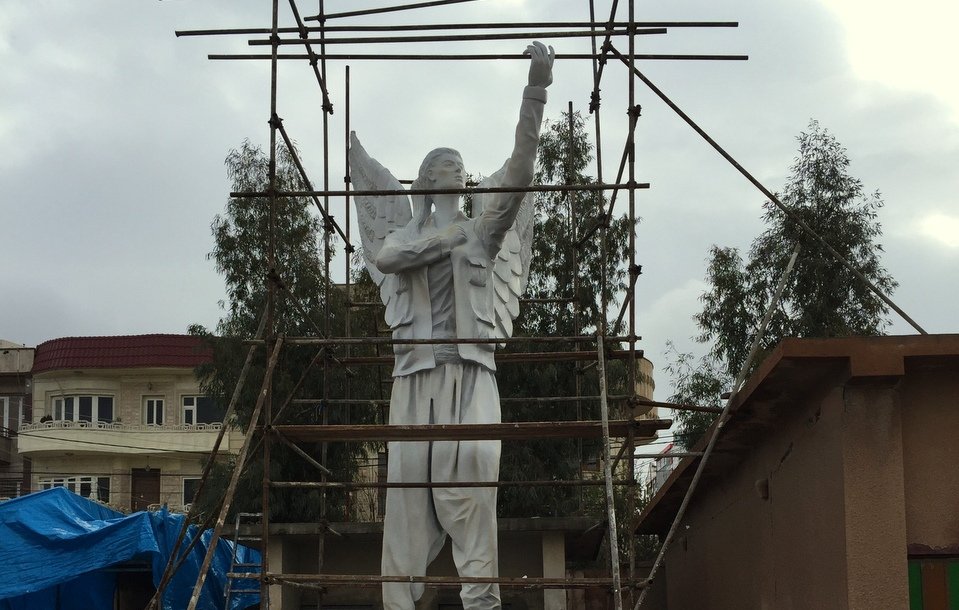 L'image montre une statue de grande taille, représentant un personnage avec des ailes, qui semble être en train d'être rénovée ou construite. Des échafaudages entourent la statue, indiquant qu'elle est encore en travaux. En arrière-plan, on peut entrevoir des bâtiments et une structure en toile bleue, suggérant une ambiance urbaine. Le ciel est nuageux, ce qui donne une atmosphère un peu mélancolique à la scène.