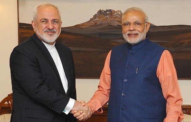 L'image montre deux hommes se serrant la main dans un cadre formel. L'un d'eux porte un costume noir et une chemise blanche, tandis que l'autre est habillé d'une veste bleue et d'une chemise orange. Ils semblent être en train d'échanger une salutation amicale, et l'arrière-plan montre une œuvre d'art représentant un paysage. L'atmosphère semble cordial et professionnelle.