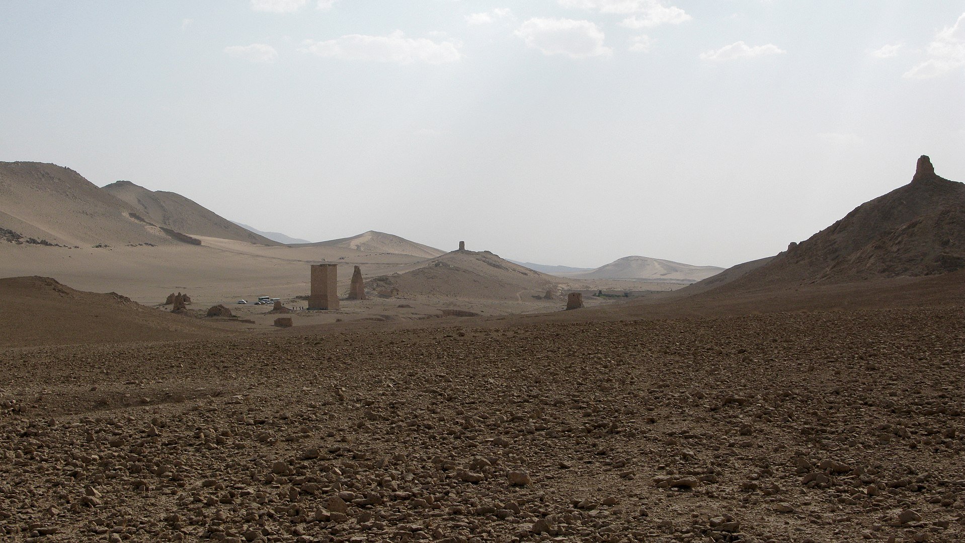 L'image montre un paysage désertique avec des collines et des formations rocheuses. Le sol est aride, parsemé de cailloux et de petites pierres. À l'horizon, on peut apercevoir quelques structures anciennes, probablement des vestiges ou des ruines, qui se détachent dans le paysage. Le ciel est clair, avec quelques nuages, et la lumière semble douce, créant une ambiance tranquille et isolée.