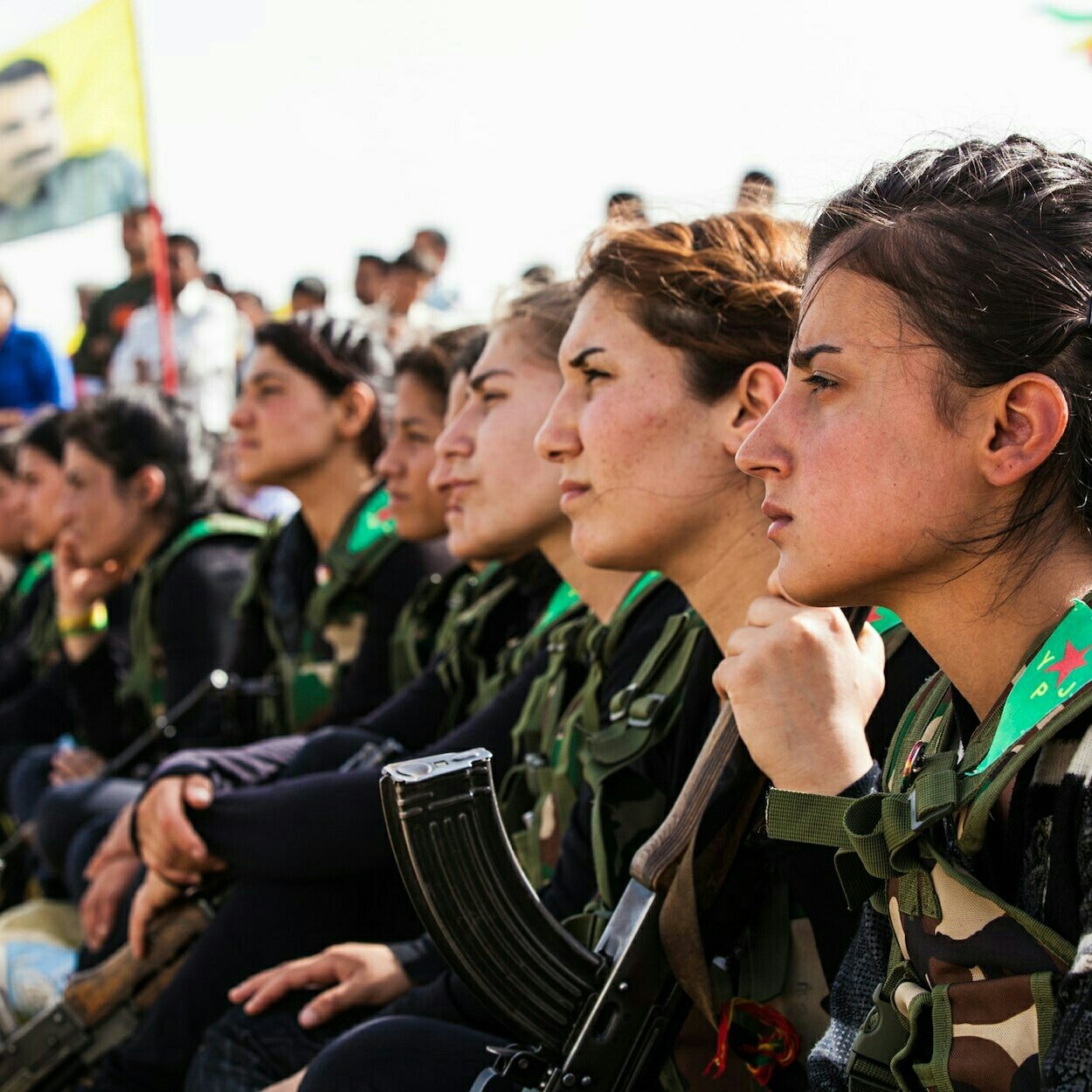 L'image montre un groupe de femmes assises en rang, portant des uniformes militaires avec des armes à leurs côtés. Elles semblent attentives et réfléchies, regardant vers l'avant. En arrière-plan, on peut apercevoir d'autres personnes réunies, ainsi qu'un drapeau symbolique. L'ambiance semble être celle d'un rassemblement ou d'une cérémonie.