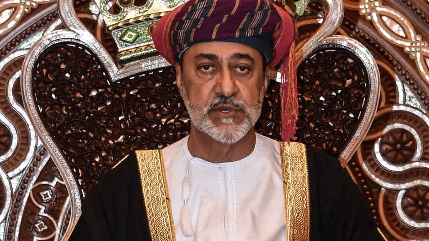 L'image montre un homme assis sur un trône orné. Il porte un costume traditionnel, comprenant une robe blanche et un turban coloré. Son expression est sérieuse et solennelle. Le fond est richement décoré avec des motifs complexes, créant une ambiance majestueuse. Les détails de la couronne et du trône mettent en valeur son statut élevé.