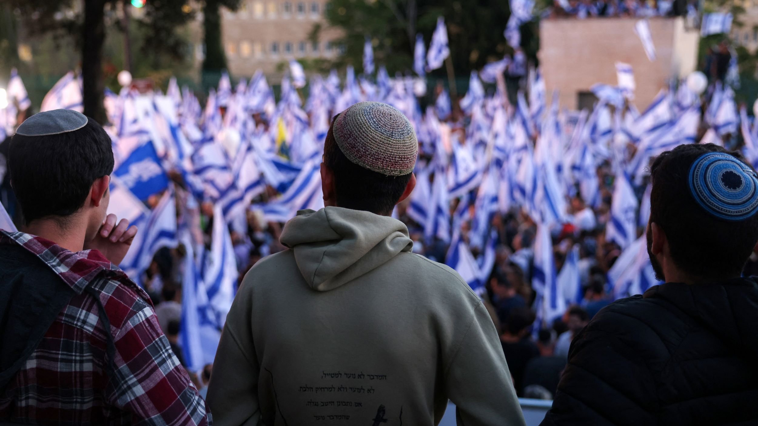 L'image montre un groupe de personnes vu de dos, regardant une manifestation ou un rassemblement. Ils portent des kippas. Au premier plan, on observe un homme en sweat à capuche, tandis que les autres sont légèrement flous. En arrière-plan, une foule dense agite des drapeaux, probablement israéliens, créant une ambiance dynamique. La scène semble se dérouler à l'extérieur, avec des arbres et des bâtiments visibles au loin.