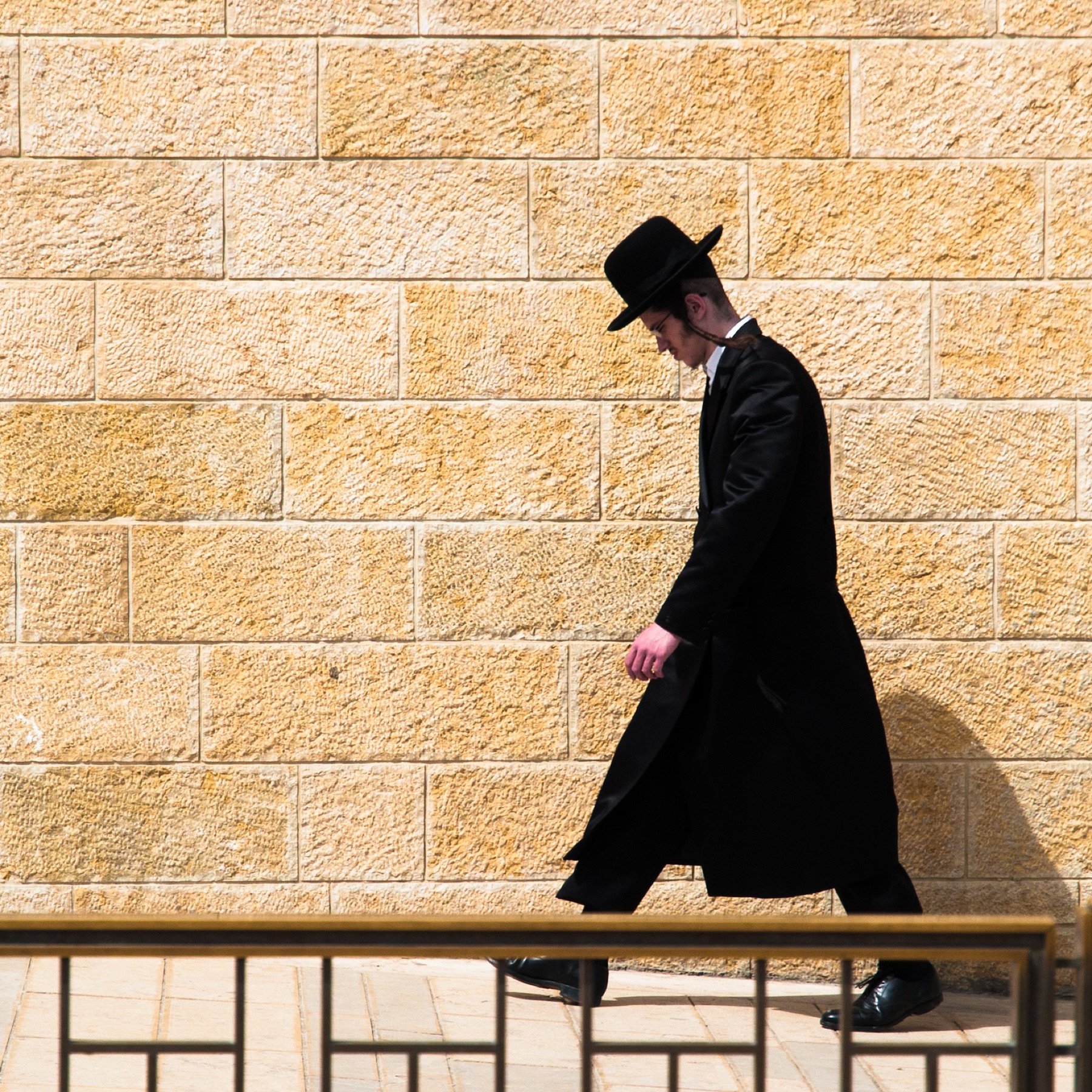 L'image montre un homme marchant le long d'un mur de pierre de couleur beige. Il est habillé d'un long manteau noir et porte un chapeau noir. Son expression est sérieuse et il semble concentré sur son chemin. L'arrière-plan est uni, ce qui met en valeur la silhouette de l'homme qui avance seul.