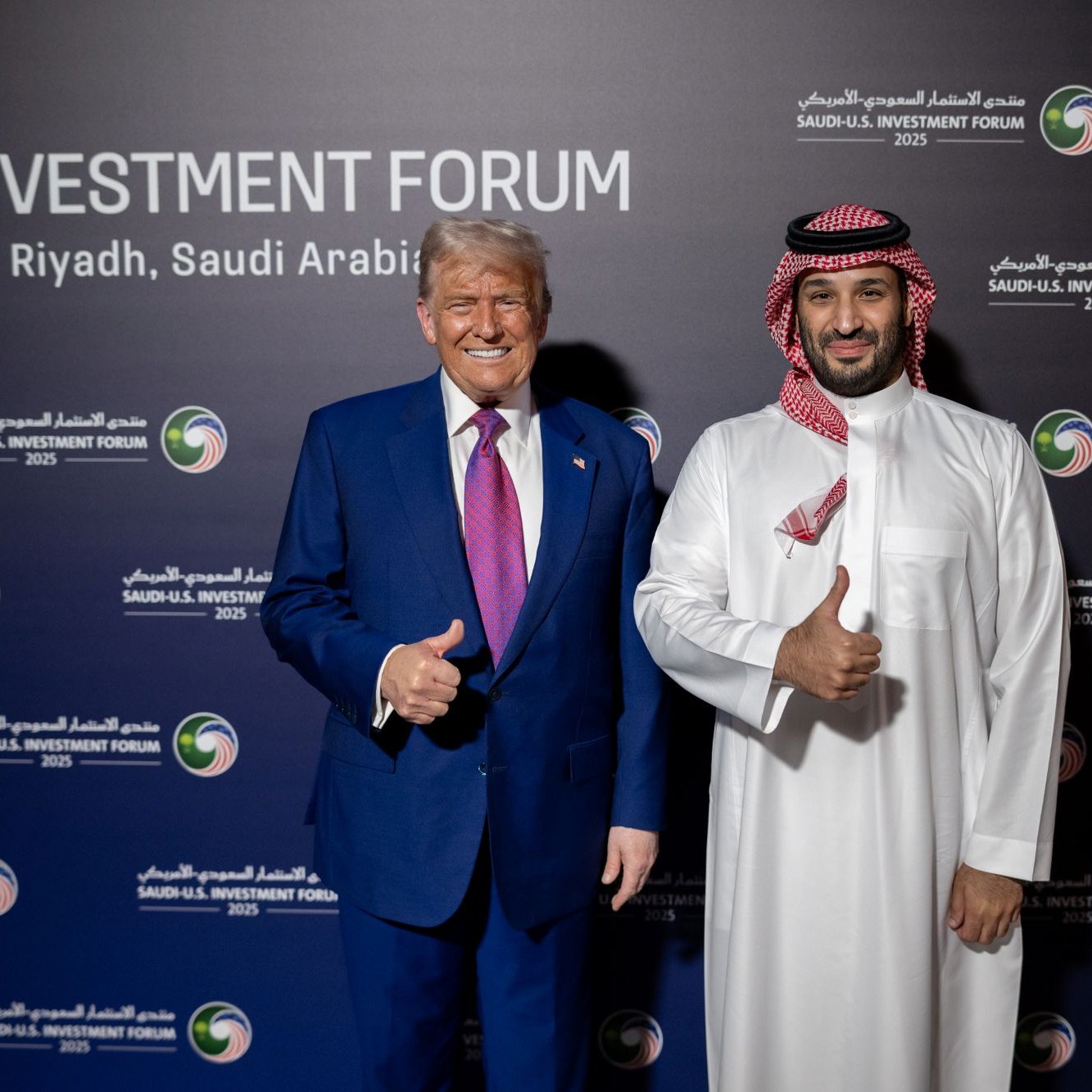 دو مرد در لباس رسمی، یکی با کت و شلوار و دیگری با لباس سنتی عربی، لبخند می‌زنند و اشاره به عالی بودن دارند.