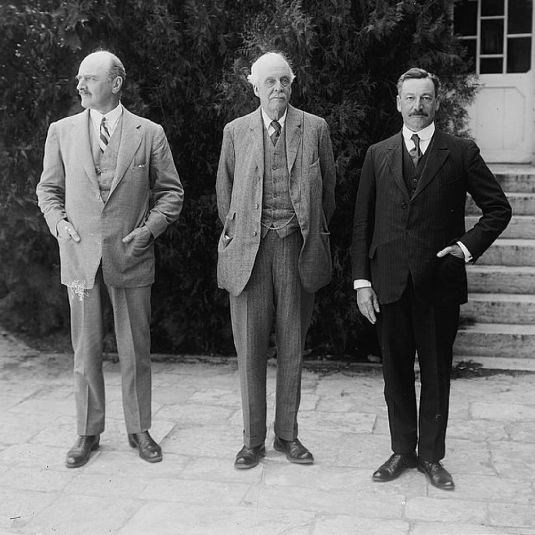 Trois hommes en costumes, debout devant un bâtiment avec des marches et des arbustes.