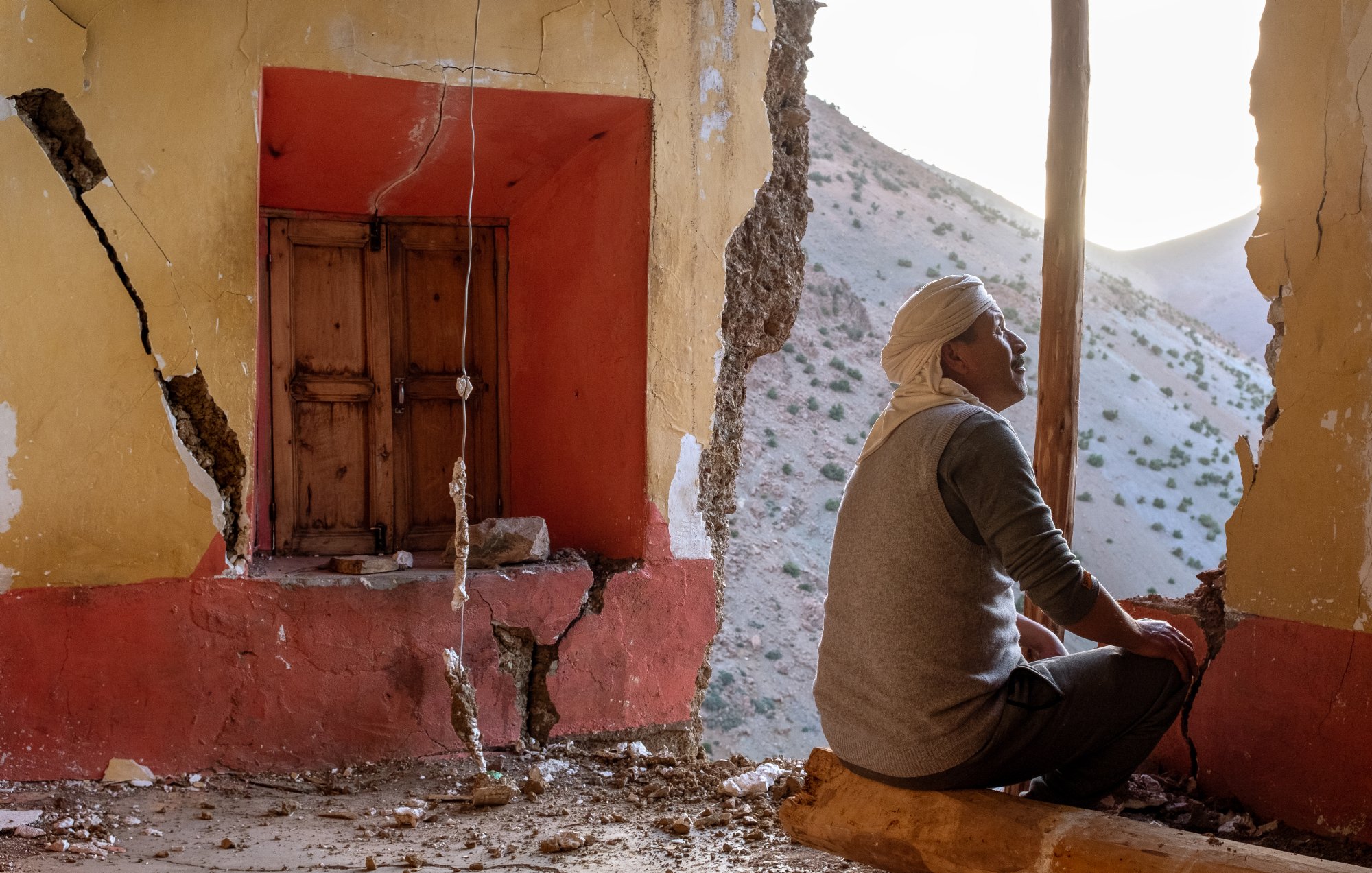L'image montre un homme assis dans une pièce aux murs endommagés, avec des fissures visibles. À sa gauche, on aperçoit une fenêtre en bois, et à droite, le mur est abîmé, laissant entrevoir l'extérieur. La lumière naturelle pénètre dans la pièce, créant une ambiance mélancolique. L'homme semble contemplatif, perdu dans ses pensées, dans un décor qui suggère l'oubli et la dégradation.