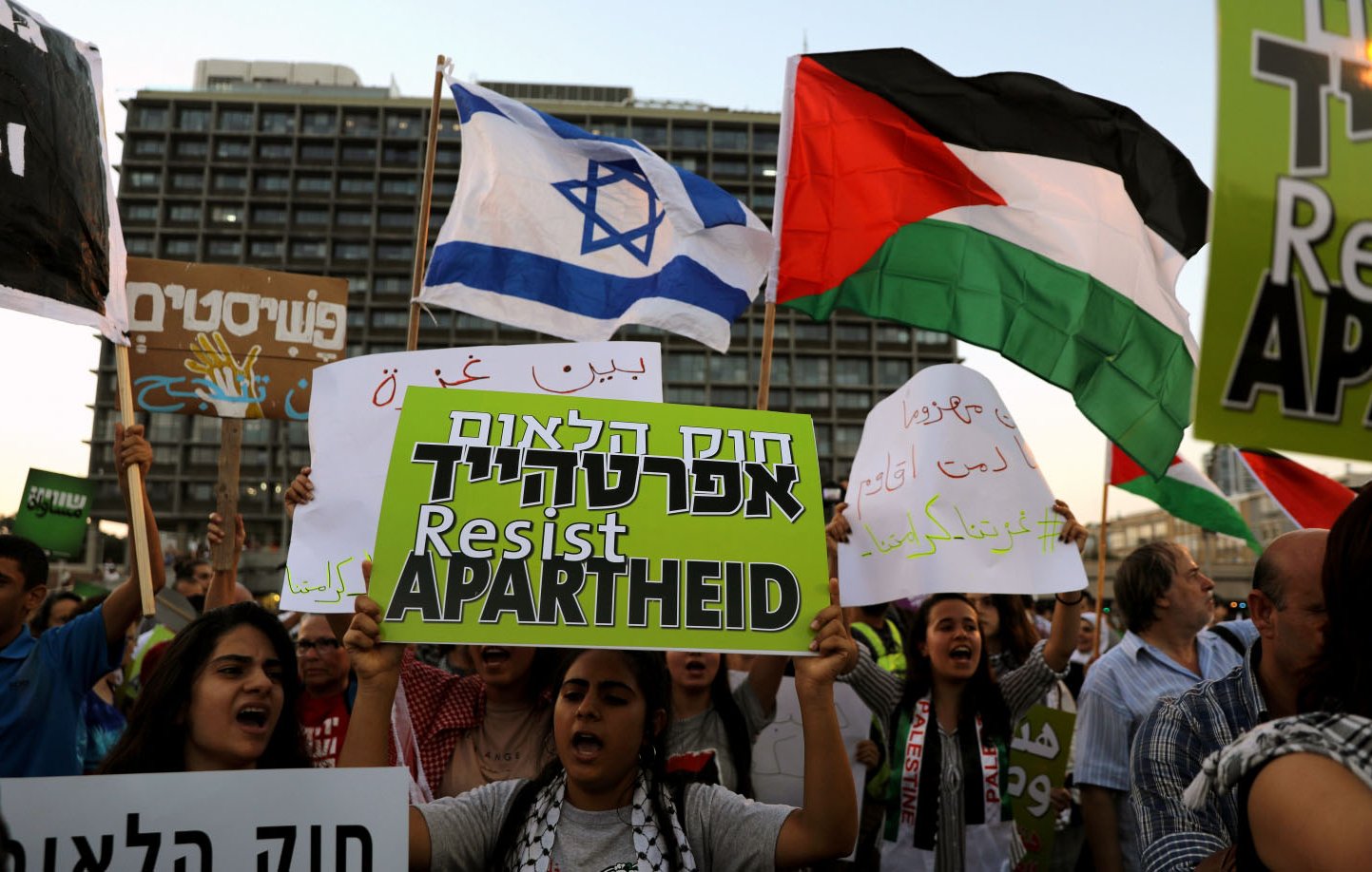L'image montre une manifestation où des gens tiennent des pancartes. Certains brandissent le drapeau israélien, tandis que d'autres agitent le drapeau palestinien. Les manifestants semblent exprimer des messages de résistance, avec des signes qui dénoncent l'apartheid. L'atmosphère est chargée d'émotion, reflet des tensions et des revendications politiques en cours.