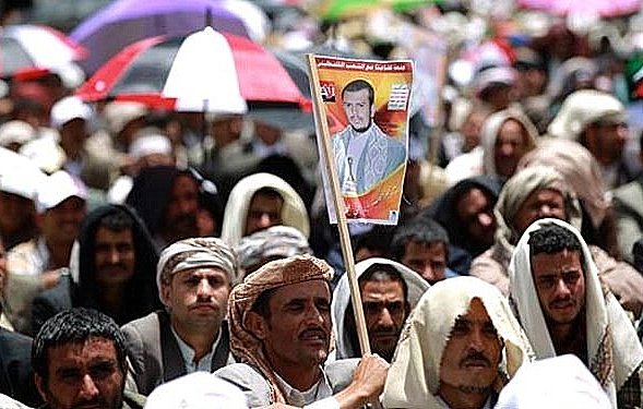 L'image montre une grande foule de personnes réunies, probablement lors d'une manifestation ou d'un rassemblement. La majorité des participants portent des vêtements traditionnels et certains sont abrités sous des parapluies de différentes couleurs. Au premier plan, un homme tient une affiche avec un portrait, ce qui suggère qu'il soutient une personnalité ou une cause spécifique. L'atmosphère semble engagée et collective.