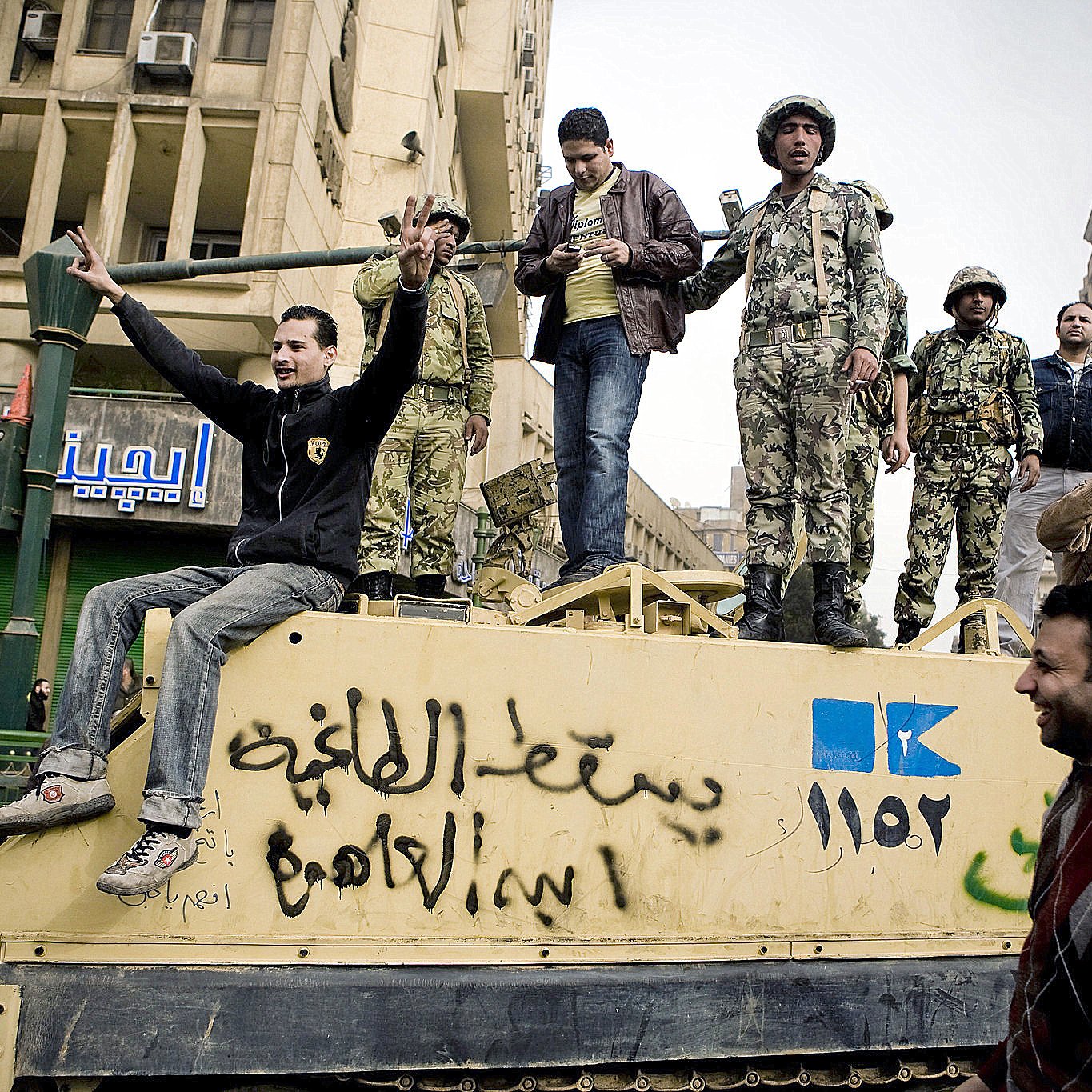 L'image montre un groupe de personnes rassemblées autour d'un véhicule militaire dans une ville. On peut voir des soldats en uniforme sur le véhicule, tandis que d'autres personnes, probablement des civils, interagissent avec eux. L'atmosphère semble tendue, mais aussi pleine d'énergie, comme si une sorte de manifestation ou de rassemblement avait lieu. L'arrière-plan montre des bâtiments urbains et des enseignes en arabe, ce qui suggère que la scène se déroule dans un pays du Moyen-Orient.