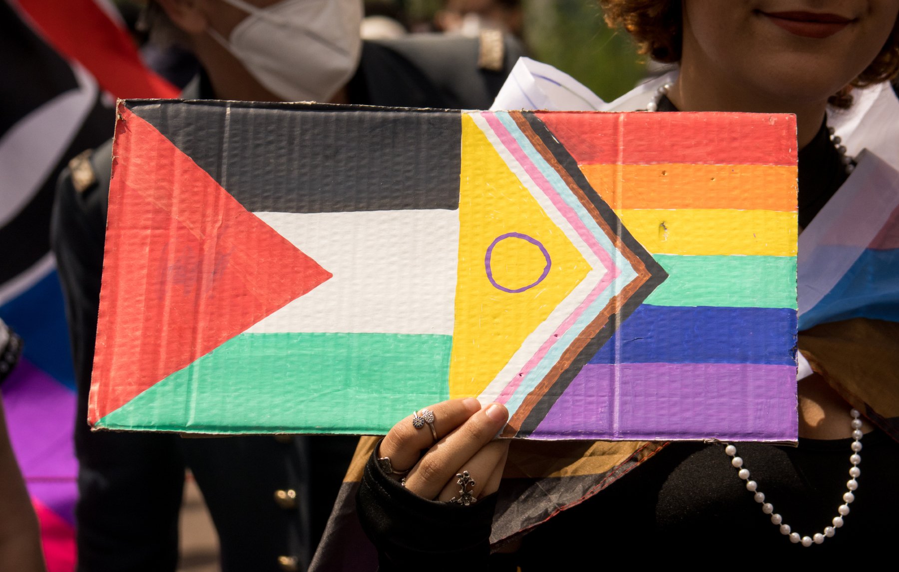 Cette image montre une personne tenant une pancarte colorée. La pancarte combine le drapeau palestinien, reconnaissable par ses bandes horizontales noire, blanche et verte avec un triangle rouge à gauche, et le drapeau Progress Pride, qui inclut les couleurs de l'arc-en-ciel ainsi que des bandes supplémentaires pour représenter la communauté transgenre et les personnes de couleur. La personne semble être en extérieur et porte une perle autour du cou ainsi que plusieurs bagues. Une autre personne portant un masque blanc est visible à l'arrière-plan.