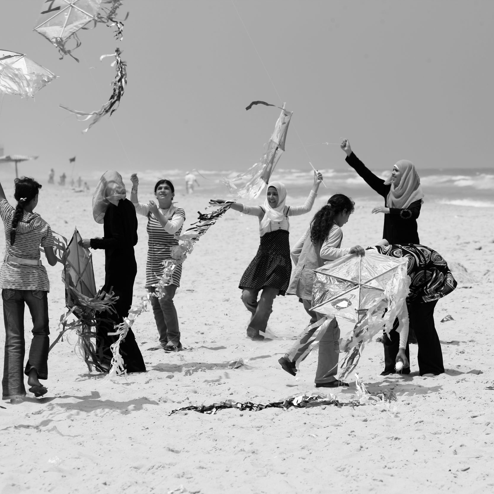 L'image montre un groupe de femmes sur une plage, s'amusant à faire voler des cerfs-volants. Certaines d'entre elles sont en train de tenir ou de manipuler des cerfs-volants, tandis que d'autres semblent rire et profiter du moment. L'environnement est ensoleillé, avec des vagues en arrière-plan. La scène dégage une atmosphère joyeuse et festive, mettant en avant des interactions sociales et des activités de plein air.