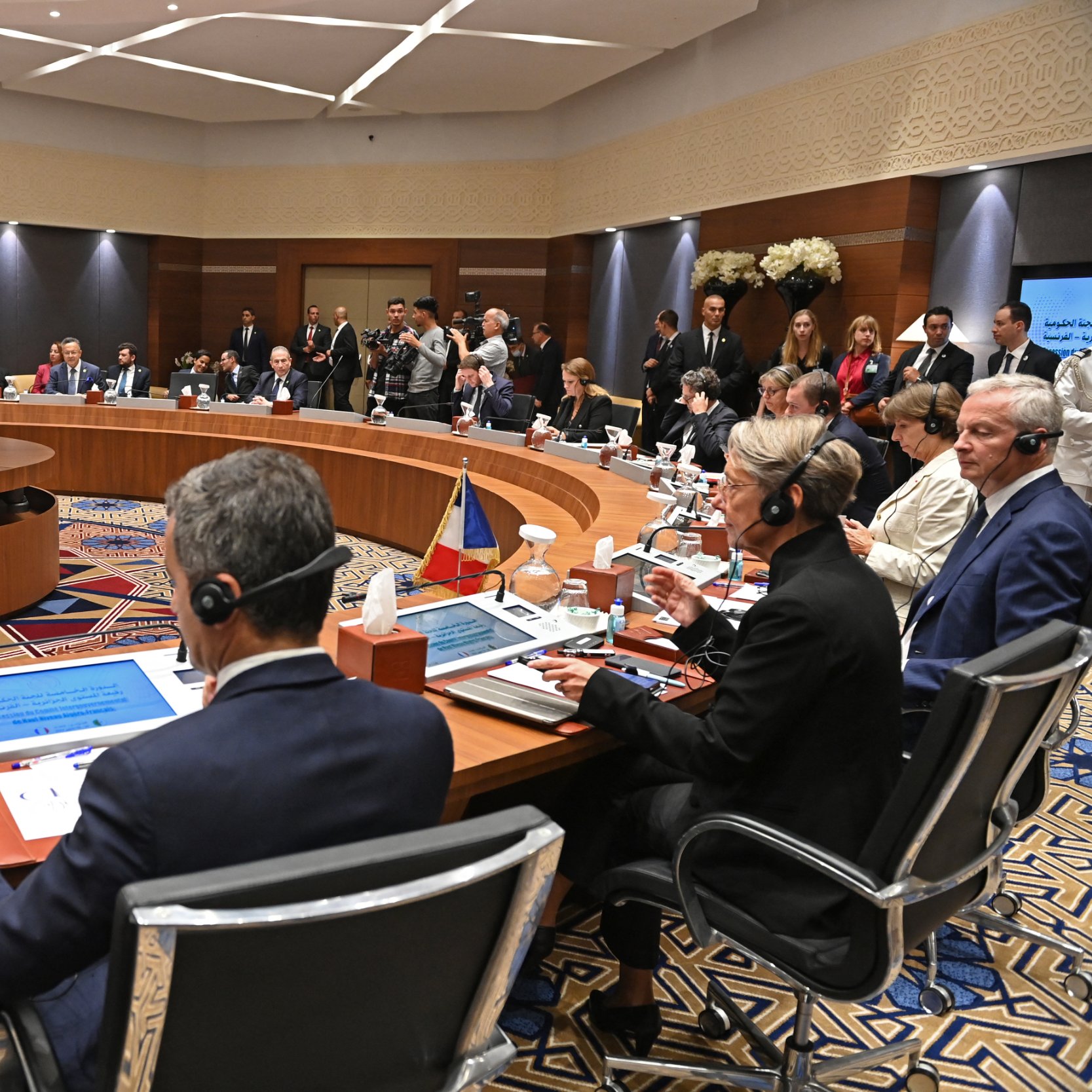 تظهر الصورة اجتماعًا يُعقَد في قاعة مؤتمرات، حيث يجلس العديد من المشاركين حول طاولة دائرية. تتواجد شاشات عرض أمام بعض الأشخاص، وتبدو القاعة مزينة ببعض الزهور. يُلاحظ وجود عدد من الحضور في الخلفية، مما يشير إلى أهمية الحدث. المشاركون يرتدون ملابس رسمية ويظهرون في لحظة من النقاش أو الاستماع.