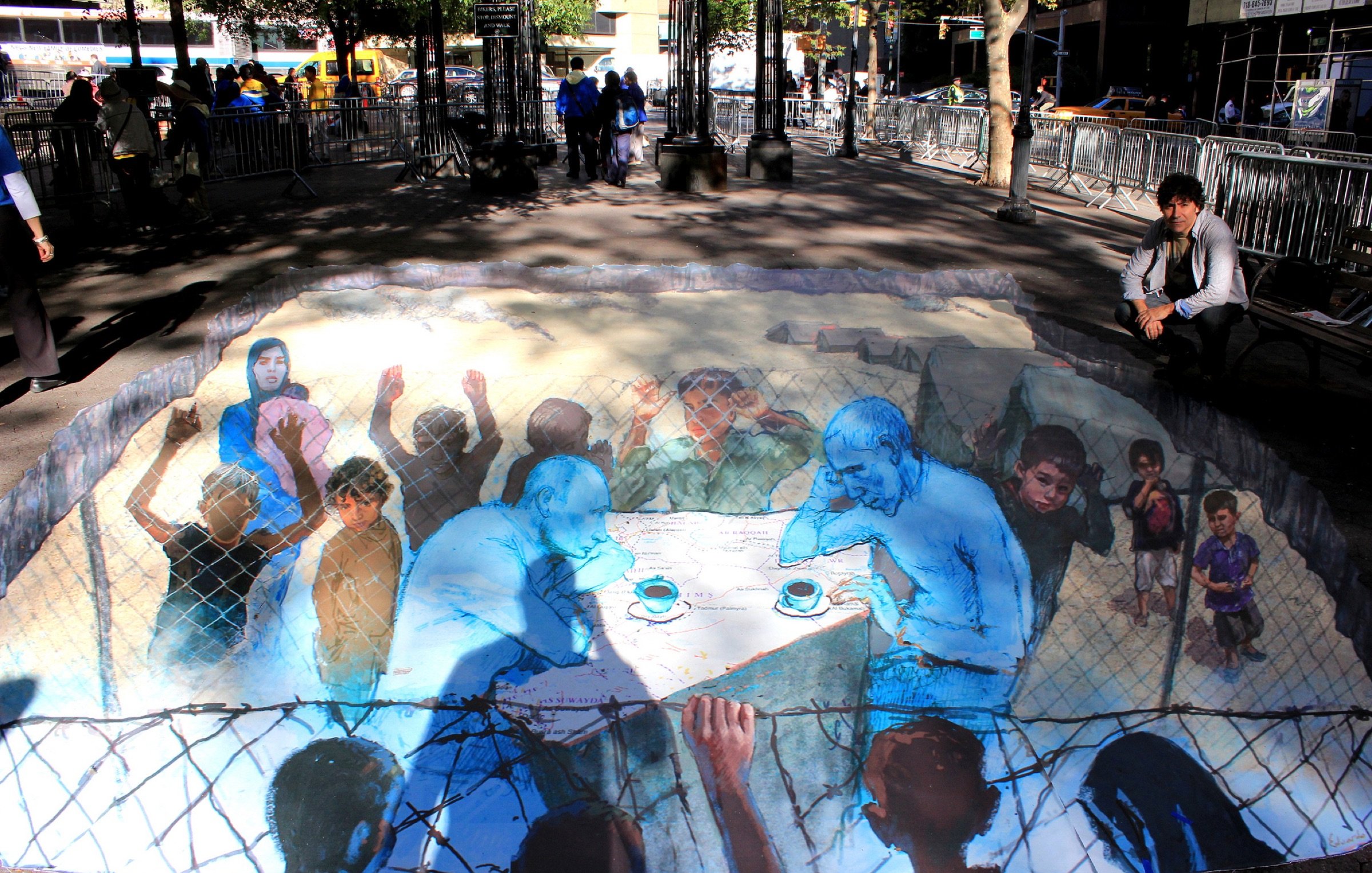 L'image montre une œuvre d'art en 3D peinte sur le sol, représentant des figures humaines engagées dans une conversation. On y aperçoit deux hommes assis à une table, entourés de scènes de détresse, symbolisant possiblement des personnes en situation de crise. Des enfants et d'autres figures semblent observer, créant un fort impact émotionnel. La couleur bleue prédomine, ajoutant à la gravité de la scène. L'environnement urbain autour de l'œuvre indique que cela se déroule dans un espace public, où des passants peuvent réfléchir sur le message véhiculé par cette peinture.