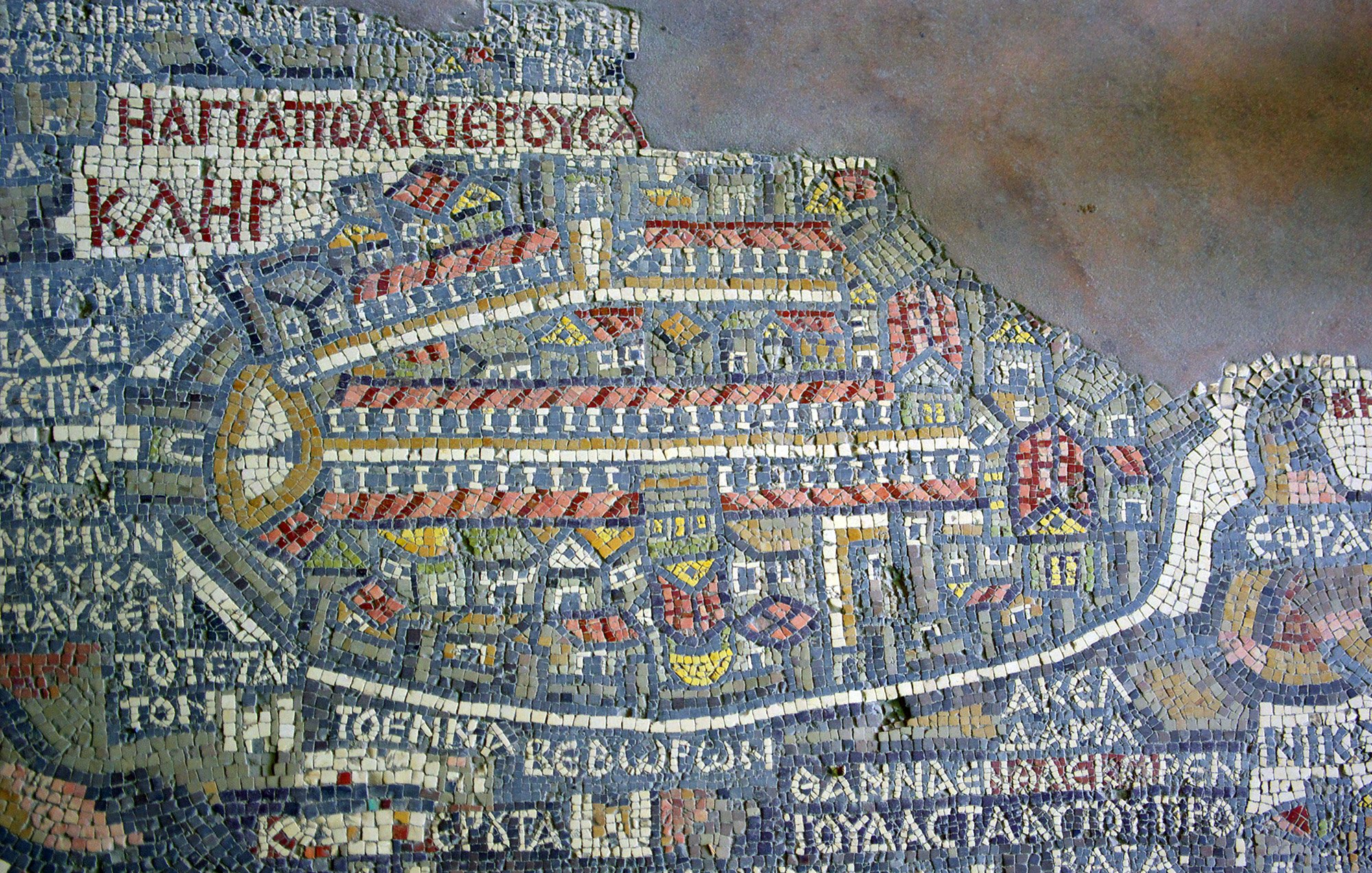 Cette image montre un ancien mappemonde représentant la ville de Jérusalem, réalisée en mosaïque. On y distingue des motifs géométriques et des bâtiments stylisés, disposés de manière à évoquer la topographie de la ville. Les inscriptions en grec ancien ajoutent des détails sur les lieux et les quartiers. La mosaïque utilise une palette de couleurs variées, créant un effet visuel riche et complexe, typique de l'art byzantin.
