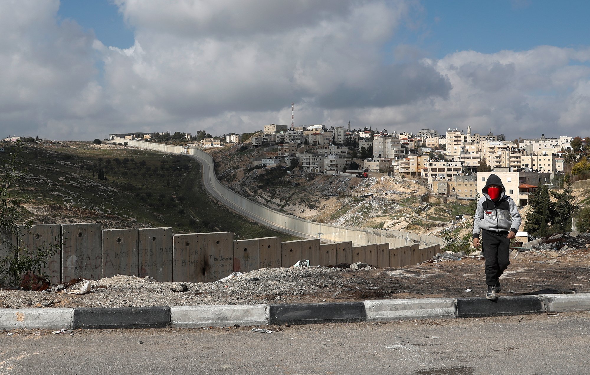 L'image montre un enfant marchant le long d'une route, avec des blocs de béton formant une barrière sur le côté. En arrière-plan, on peut voir une colline surmontée de bâtiments, indiquant une zone urbaine. Le ciel est partiellement nuageux, et le paysage semble aride et rocailleux. L'enfant porte une capuche et un masque, ajoutant une dimension de mystère à la scène. La barrière suggère des thèmes de division et de conflit.