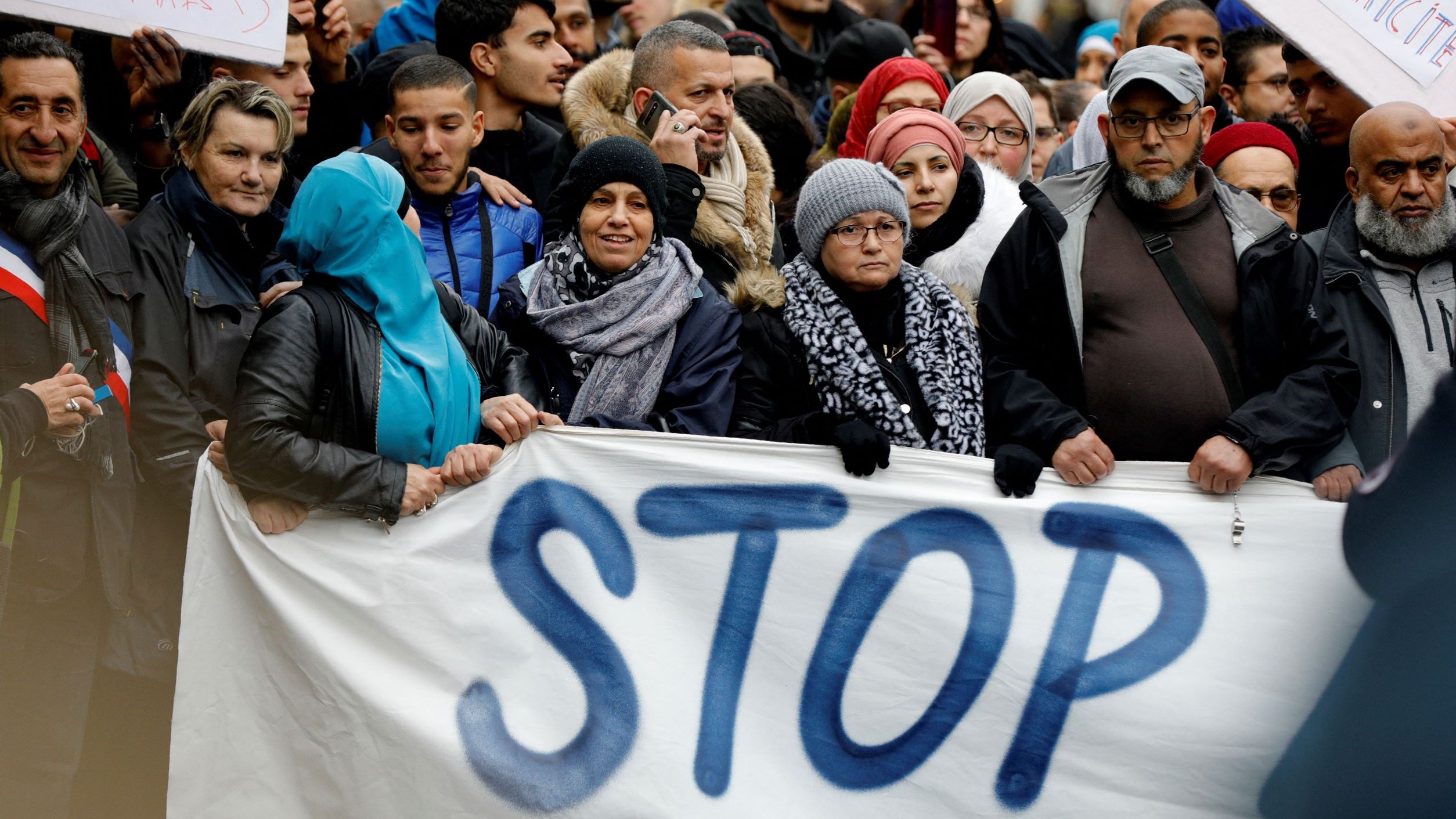 L'image montre une foule de personnes rassemblées pour une manifestation. Elles tiennent des pancartes, dont l'une affiche le mot "STOP" écrit en grandes lettres. Les participants portent des vêtements variés, certains ayant des écharpes ou des chapeaux, ce qui suggère qu'il pourrait faire frais. L'atmosphère semble engagée, avec des visages sérieux ou déterminés, reflétant un fort sentiment de solidarité pour la cause qu'ils soutiennent.
