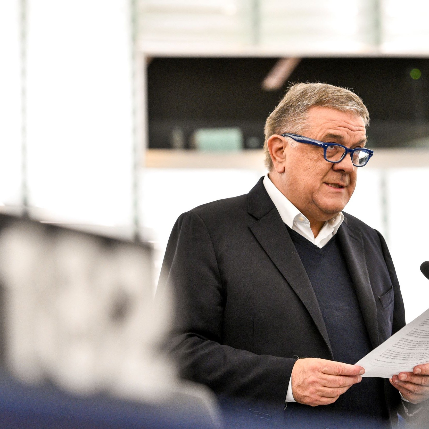 Nell'immagine si vede un uomo che parla a un microfono, probabilmente durante una sessione al Parlamento Europeo. Indossa un blazer scuro e occhiali blu. Tiene in mano un foglio, probabilmente un discorso o una relazione, e sembra essere concentrato mentre si rivolge al pubblico. Sullo sfondo si intravedono alcune strutture dell'ambiente parlamentare.