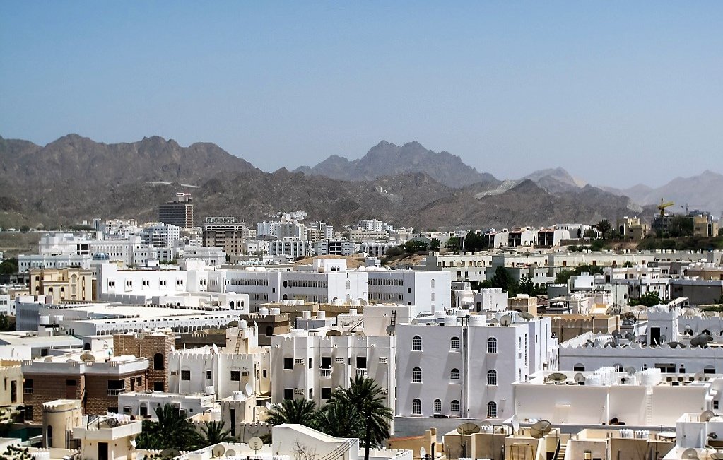 الصورة تُظهر مشهداً لمدينة محاطة بالجبال، حيث تتداخل المباني البيضاء مع بعض اللمسات الحديثة. تمتاز rooftops بأسقفها المسطحة، بينما تظهر في الخلفية الجبال الشاهقة التي تضفي جمالاً على المنظر. السماء نقية وزرقاء، مما يضيف إحساساً بالصفاء والهدوء للمنطقة.