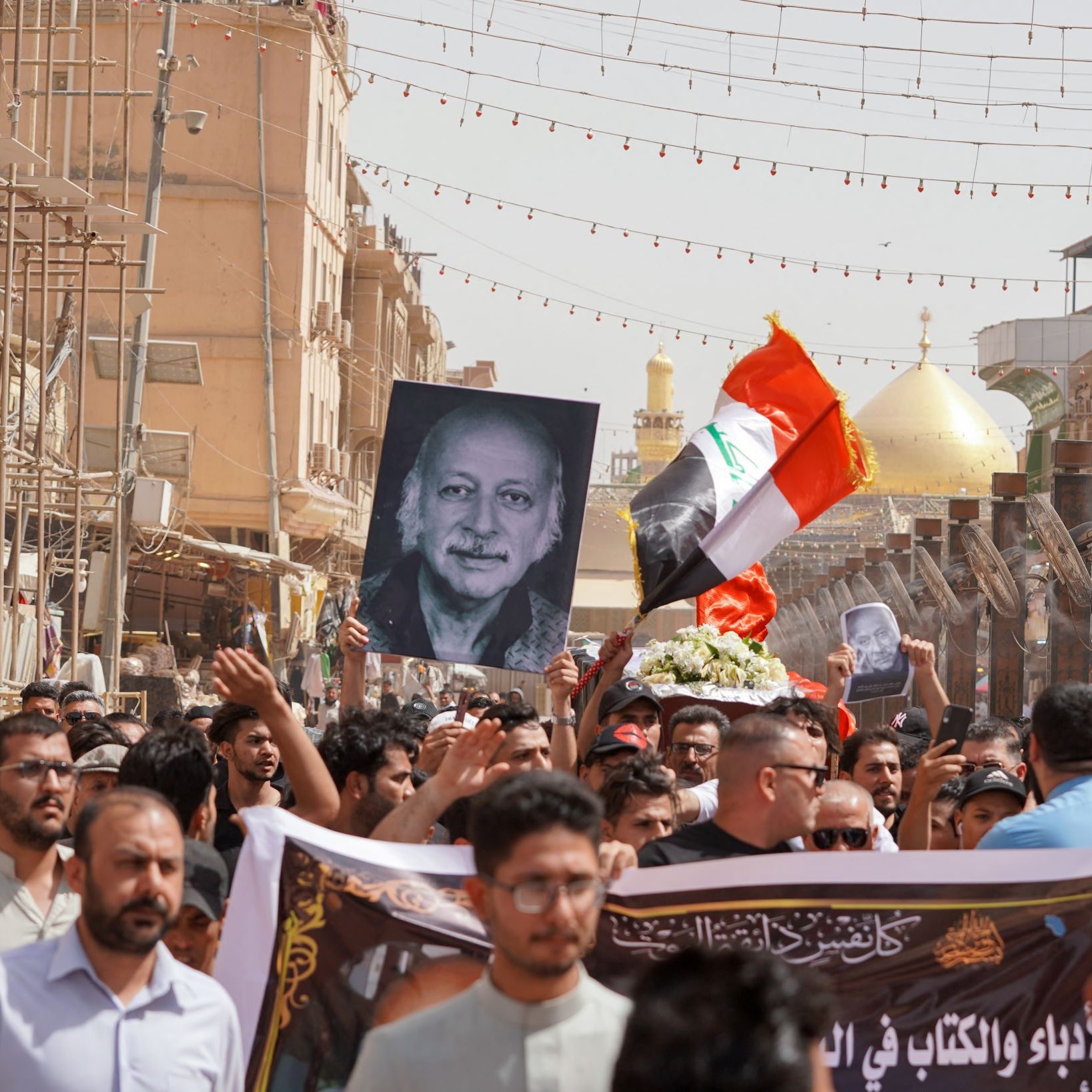 L'immagine mostra una folla di persone durante una manifestazione. Molti manifestanti tengono in alto una grande fotografia di un uomo, insieme a una bandiera irachena. Sullo sfondo si possono vedere elementi architettonici e due cupole dorate, che suggeriscono la presenza di un luogo sacro o importante. L'atmosfera sembra intensa e significativa, con i partecipanti che mostrano il loro sostegno o commemorazione attraverso slogan e simboli.