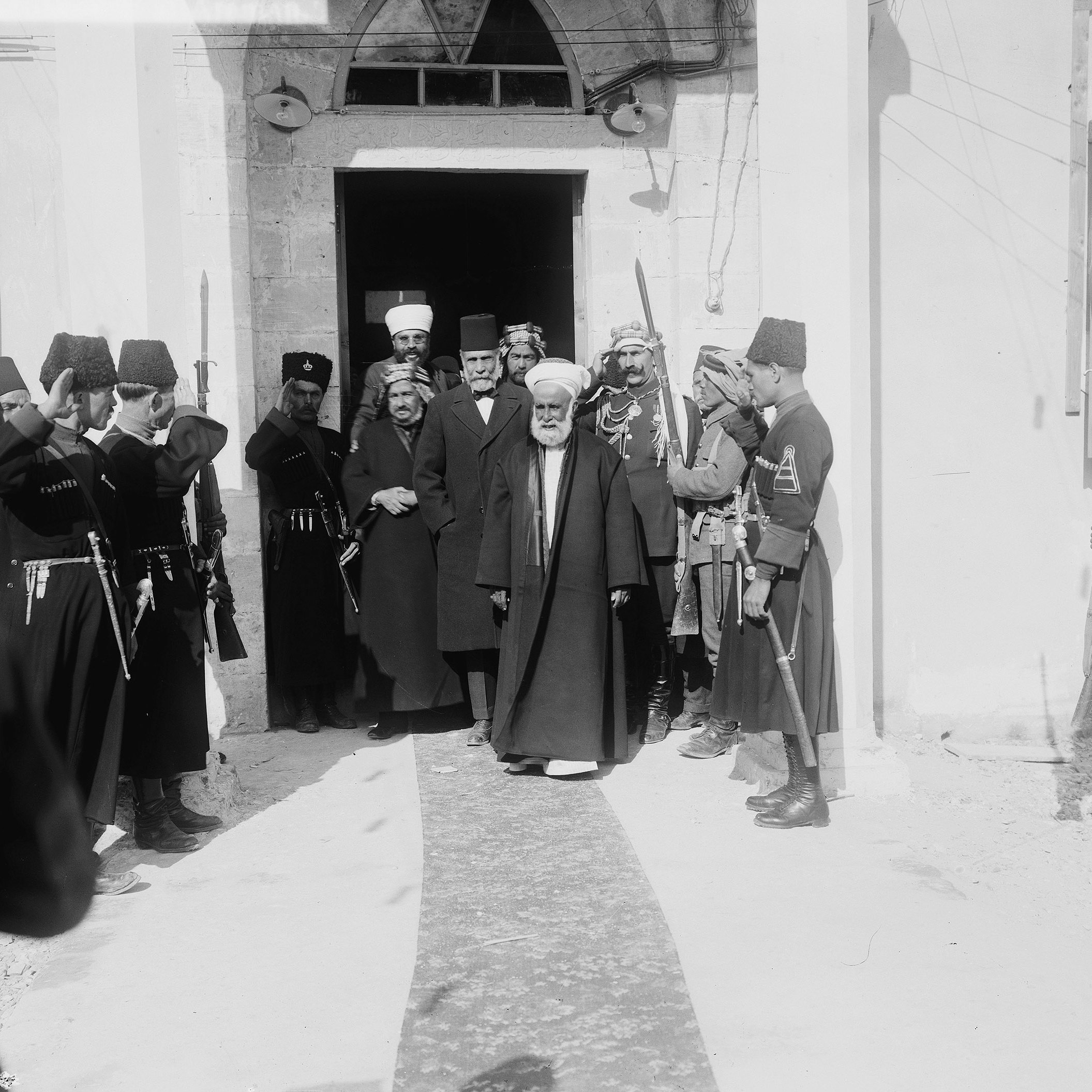 The image depicts a formal gathering outside a building, likely a historical site. A group of men dressed in traditional attire, including some wearing fezes and military uniforms, are seen saluting. In the center, an older man in a long robe and head covering is walking out of the entrance, suggesting he holds a position of significance or authority. The surrounding individuals, including soldiers and other men in formal dress, appear to be honoring or welcoming him. The scene conveys a sense of respect and formality typical of ceremonial occasions. The architecture hints at a historical and possibly Middle Eastern context.
