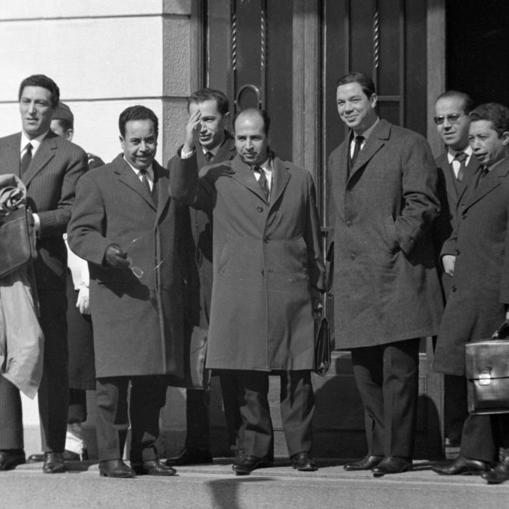 L'image montre un groupe de plusieurs hommes en costume, se tenant à l'extérieur d'un bâtiment. Ils portent des manteaux d'hiver, indiquant que la scène se déroule probablement par temps frais. L'un des hommes, au centre, semble être en train de saluer ou de parler avec enthousiasme. Les autres hommes affichent des expressions neutres ou souriantes. L'environnement est urbain, avec des détails architecturaux en arrière-plan.