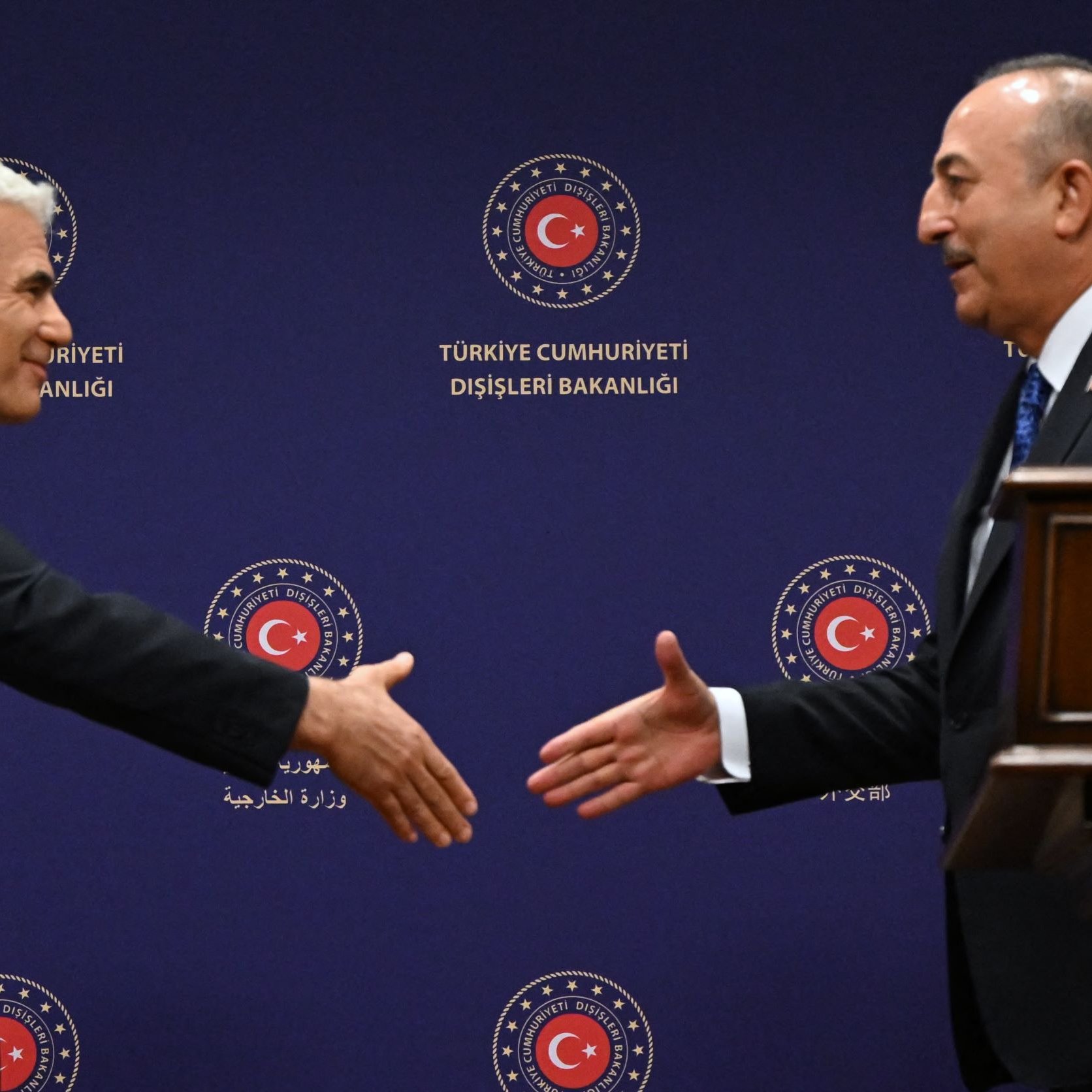 The image depicts two men engaged in a handshake, likely at a diplomatic or formal meeting. They are standing on either side of a wooden podium, with a background featuring the logo of the Turkish Ministry of Foreign Affairs. One man appears to be offering his hand in a gesture of greeting, while the other is responding. Both are dressed in formal attire, suggesting a professional context.