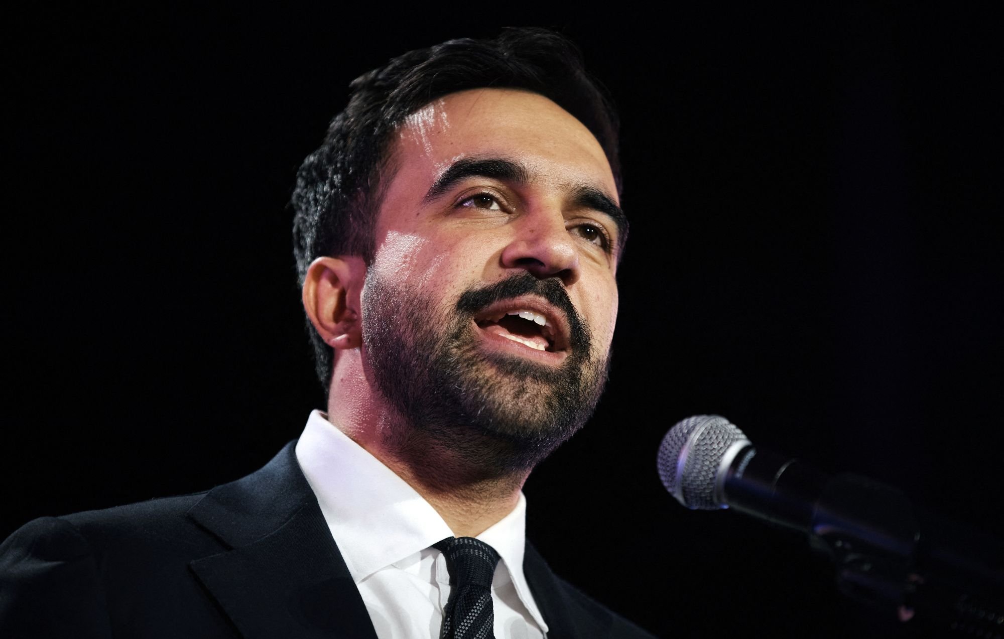 Homme en costume noir, parlant au micro avec une expression engagée.