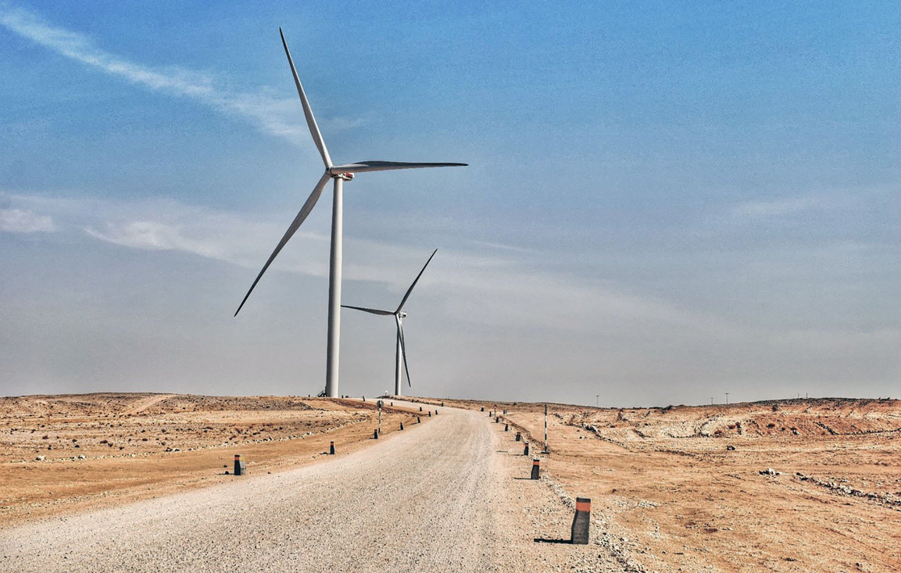 La imagen muestra un paisaje árido y desértico con un camino de grava que se extiende hacia el horizonte. A ambos lados del camino, se pueden ver aerogeneradores, que son grandes turbinas eólicas que aprovechan la energía del viento. El cielo está despejado y presenta un tono azul, lo que sugiere un día soleado. La escena transmite una sensación de tranquilidad y sostenibilidad, con la tecnología eólica destacando en un entorno natural.