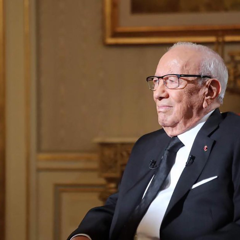 تظهر الصورة شخصًا مسنًا يجلس على كرسي في غرفة ذات ديكور فخم. يرتدي بدلة رسمية سوداء مع قميص أبيض وربطة عنق داكنة. يضع نظارات على عينيه ويبدو عليه التفكير والتركيز. خلفه توجد جدران مزخرفة وأمكنة تزينها الأعمال الفنية، مما يضيف لمسة من الأناقة إلى المشهد.