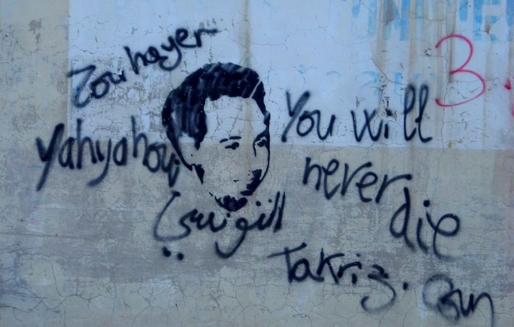 L'image représente un graffiti sur un mur. On y voit un visage stylisé, probablement d'une personne, entouré de mots écrits à la main. En haut, il y a le texte en arabe "Zou haya" et "Yahya", et en bas, le message en anglais "You will never die", accompagné du mot "Takríz" et du mot "Gun". L'ensemble dégage un sentiment de mémoire ou d'hommage, avec un mélange de langues.
