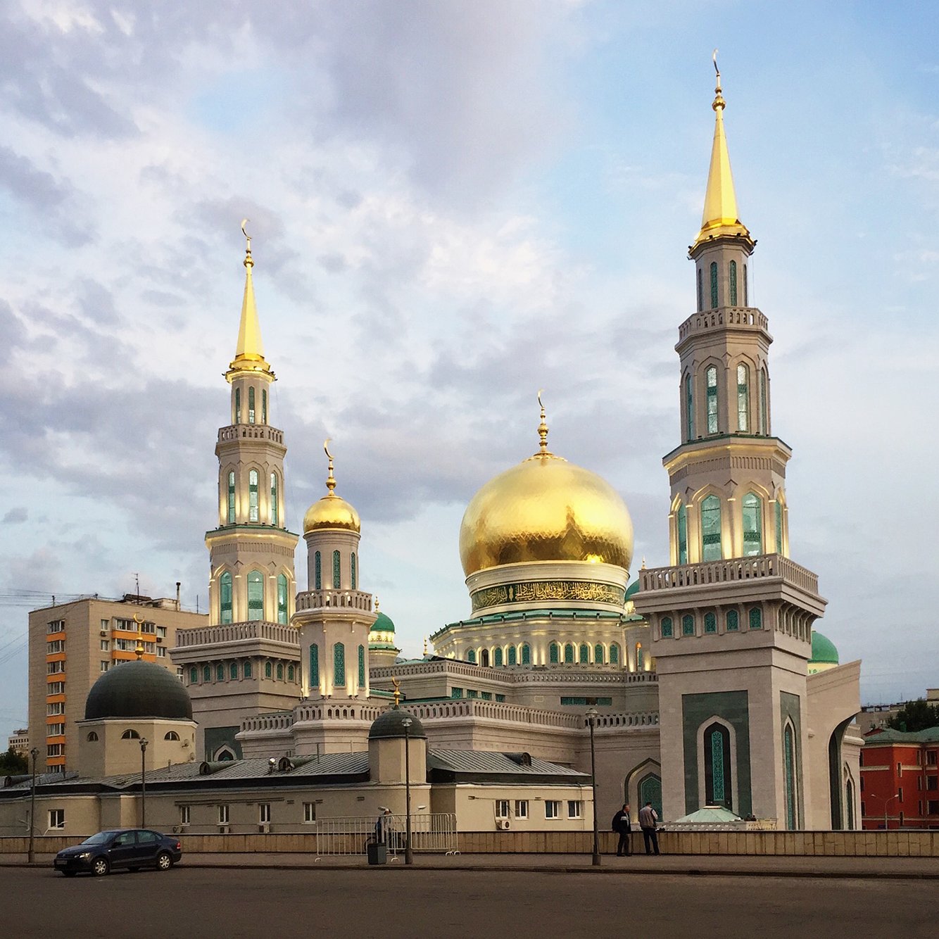 L'image montre une mosquée impressionnante avec des coupoles dorées et plusieurs minarets. Les structures sont ornées de détails architecturaux colorés, et le ciel en arrière-plan est légèrement nuageux, créant une ambiance tranquille. La mosquée se dresse majestueusement en milieu de ville, entourée d'immeubles modernes.