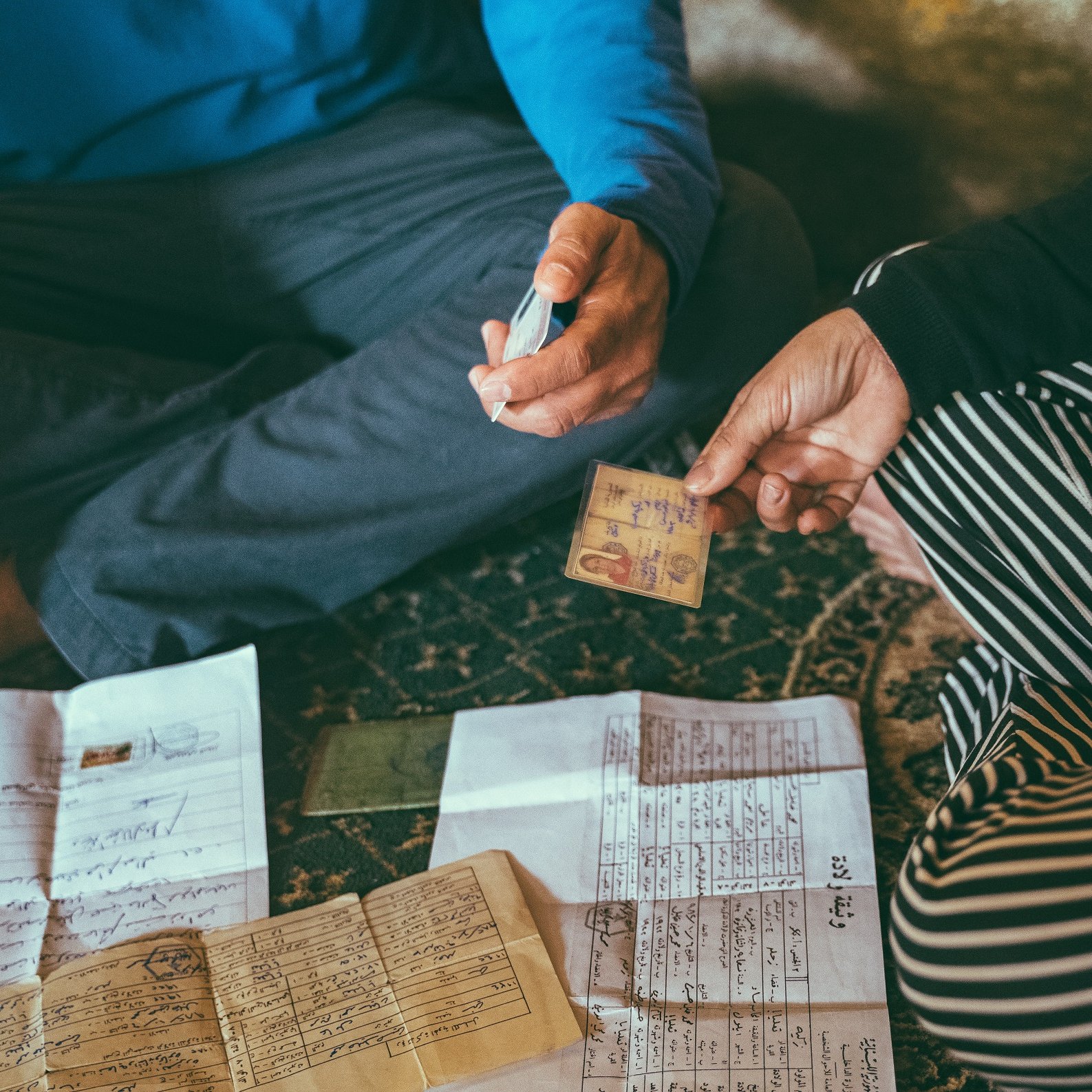 L'image montre deux personnes assises par terre, entourées de divers documents étalés devant elles. L'une des personnes tient une carte d'identité ou un document similaire, tandis que l'autre semble utiliser un marqueur pour écrire ou annoter quelque chose. Les documents sur le sol sont variés, comprenant des papiers manuscrits et des feuilles à imprimer. L'environnement semble intime et informel, suggérant une discussion importante ou une transaction en cours.