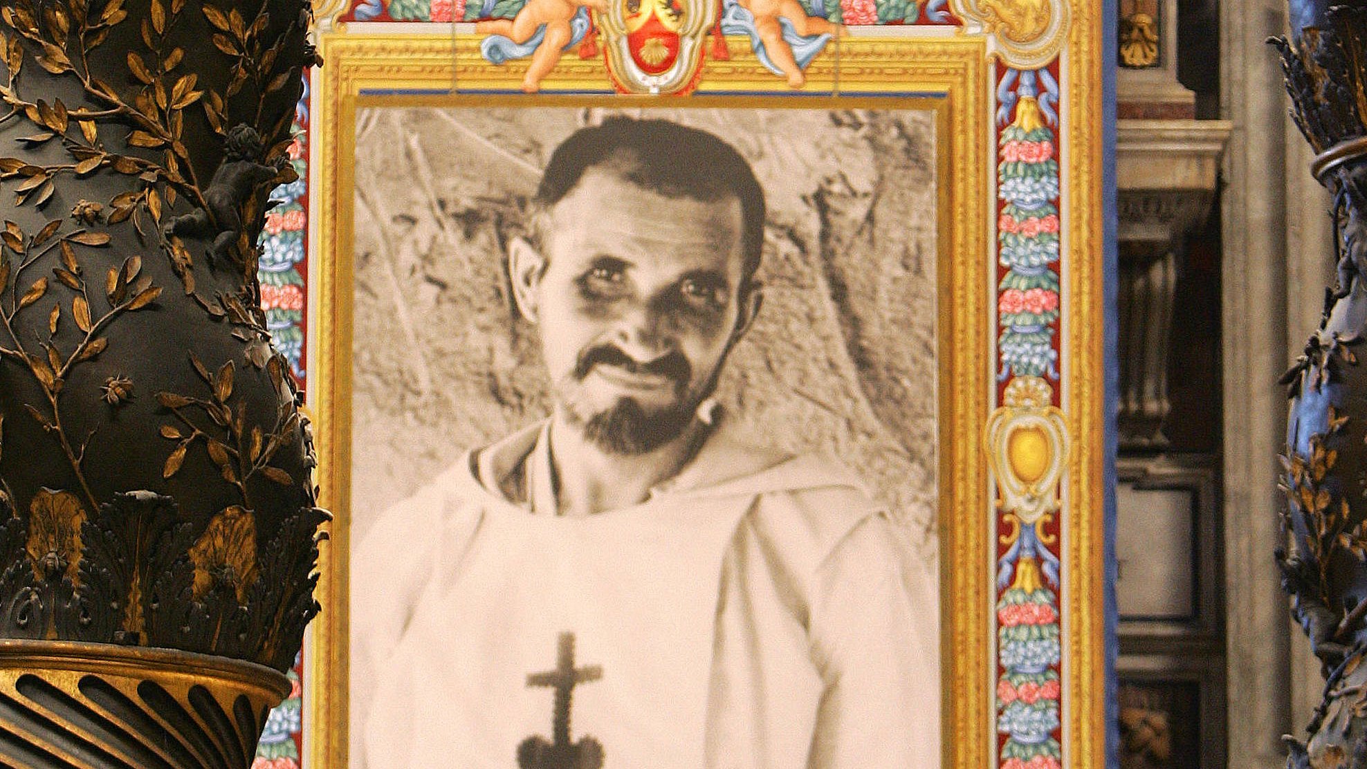 L'image montre un intérieur majestueux, probablement dans une église ou une cathédrale, avec des colonnes ornées de motifs dorés et des détails complexes. Au centre, il y a un grand portrait en noir et blanc d'un homme, qui semble vêtu d'une robe religieuse, encadré de couleurs vives et de décorations. Ce cadre attire l'œil et contraste avec la richesse des ornements de l'architecture environnante. Les murs et les éléments de la pièce évoquent une atmosphère sacrée et historique.