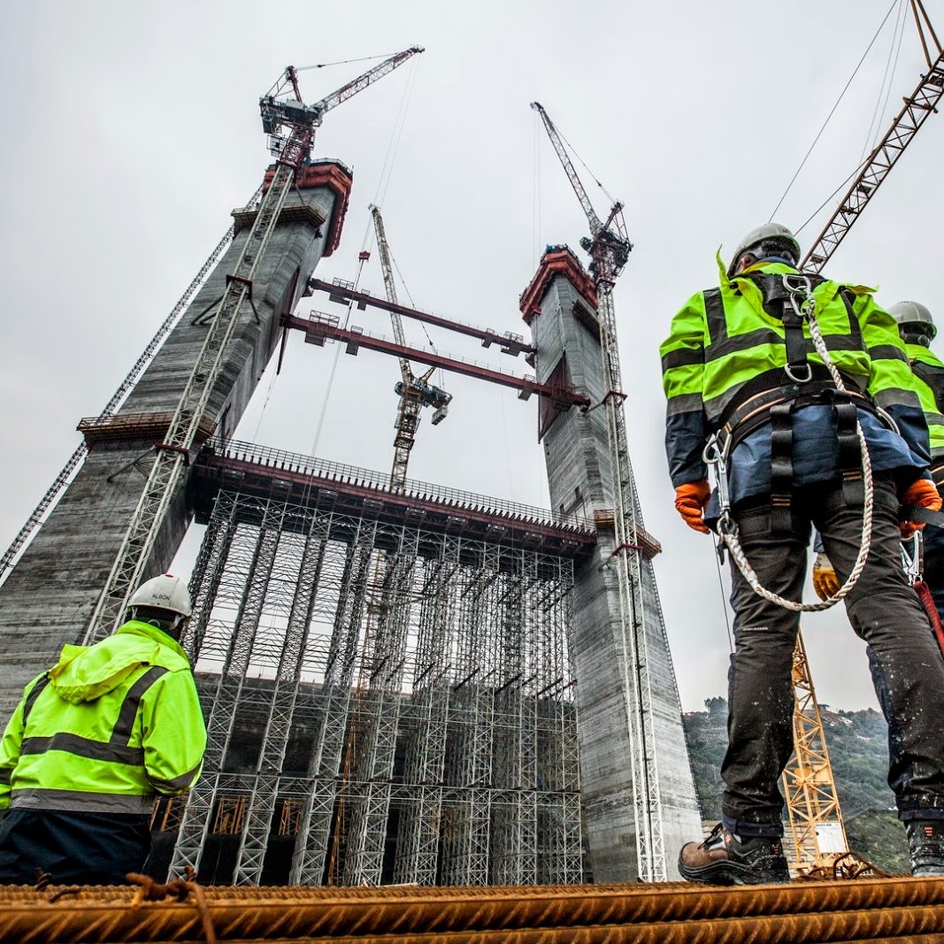 L'image montre une scène de construction avec plusieurs travailleurs vigilants observant une structure imposante en cours d'édification. Les ouvriers portent des vestes de sécurité à rayures et des casques, et sont accrochés avec des harnais de sécurité. En arrière-plan, on peut voir des grues et des éléments de construction en cours d'assemblage, tandis que le ciel est nuageux, suggérant une ambiance de travail intense sur le chantier.