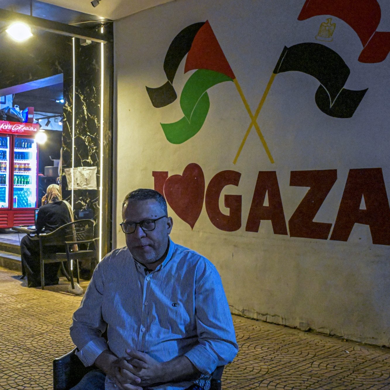 Un homme assis, devant un mural "I ❤️ GAZA", avec des drapeaux palestiniens en arrière-plan.