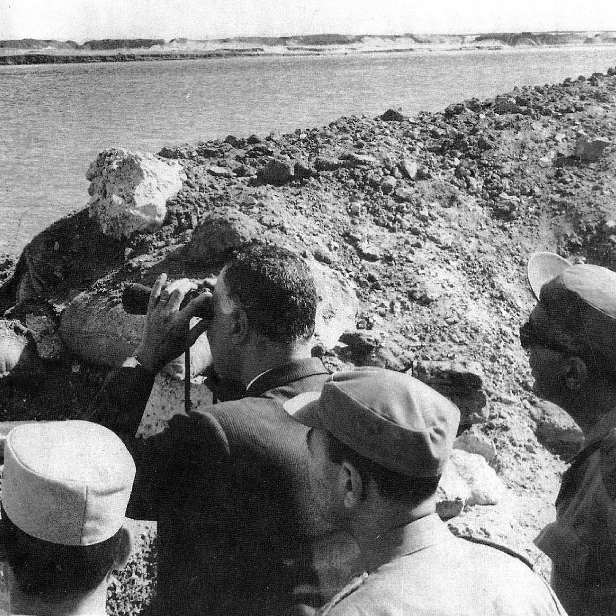 تظهر في الصورة مجموعة من الأشخاص يقفون على حافة منطقة مائية، يراقبون شيئًا ما باستخدام المنظار. يبدو أن الشخص الموجود في المقدمة مشغول بالنظر من خلال المنظار، بينما يراقبه الآخرون من حوله. الخلفية تتكون من تربة وعناصر طبيعية، مما يوحي بأن الصورة قد التُقطت في موقع استراتيجي أو أثناء زيارة ميدانية.