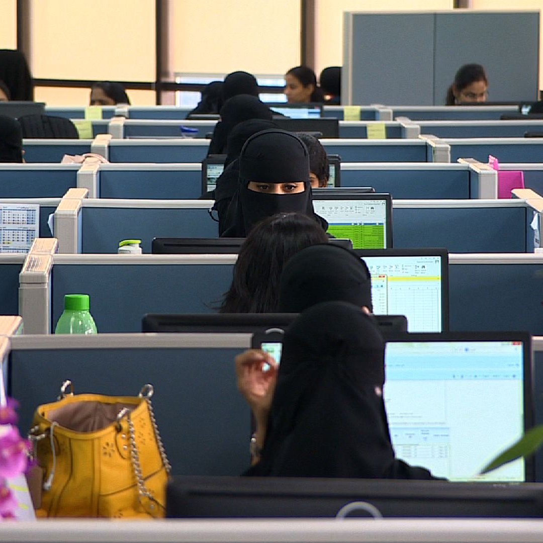 L'image montre un espace de bureau moderne, avec plusieurs employés assis à leurs bureaux, chacun devant un ordinateur. La plupart des femmes portent des vêtements noirs, y compris des hijabs ou des niqabs, ce qui leur donne une apparence uniforme. On peut voir des écrans d'ordinateur affichant des tableaux ou des données, ainsi que quelques plantes décoratives et des objets sur les bureaux. L'ambiance semble professionnelle et organisée, avec un grand nombre de personnes travaillant dans cet environnement administratif.