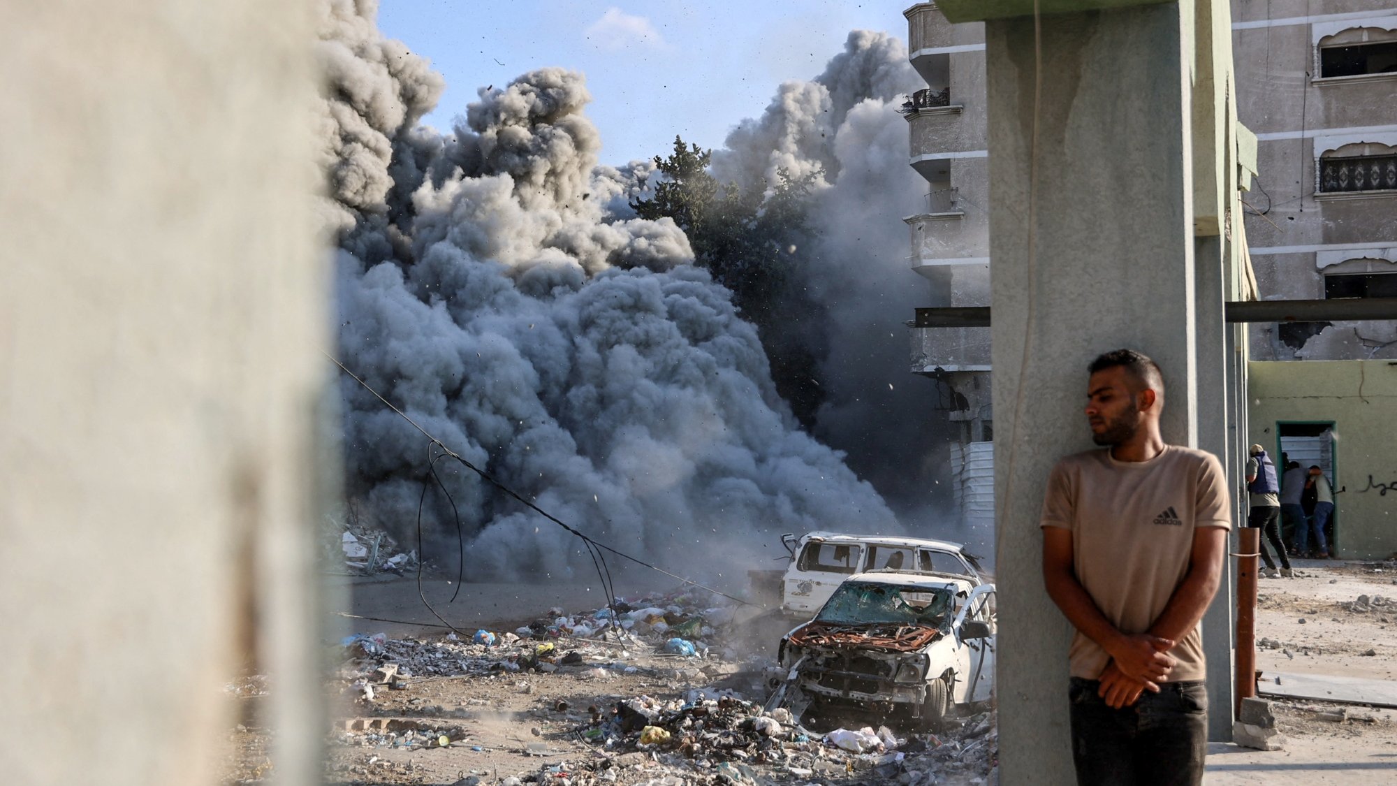 Destruction urbaine avec de la fumée, un homme adossé à un pilier près d'une voiture endommagée.