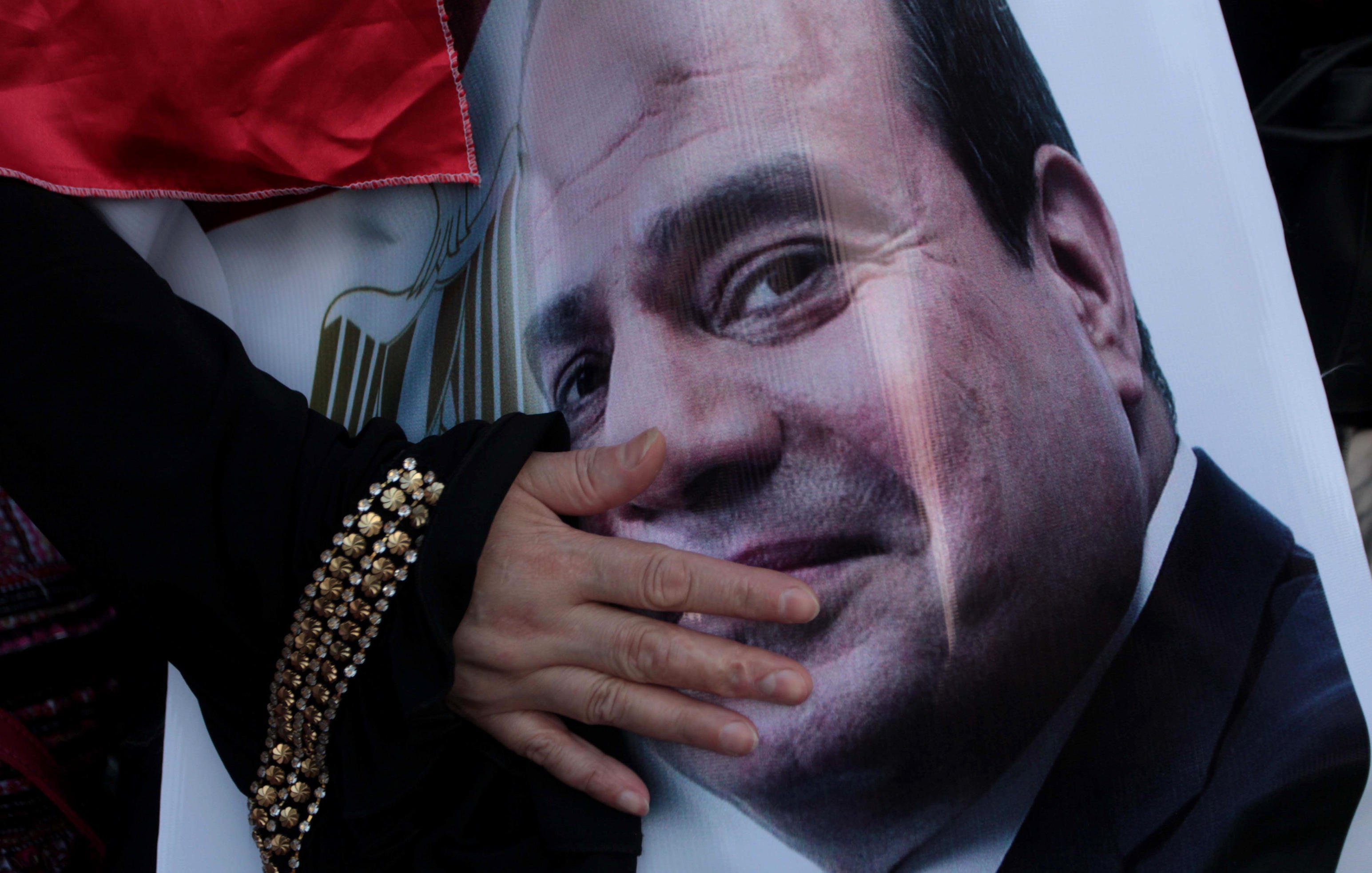 تظهر في الصورة يد تشير إلى صورة كبيرة لوجه شخص معروف، حيث توجد تعبيرات تدل على الارتياح والابتسامة. اليد تبدو وكأنها تعبر عن التأييد أو الولاء، بينما الخلفية تحتوي على ألوان تتضمن الأحمر، مما قد يشير إلى جو من الاحتفال أو التعبير عن الدعم.